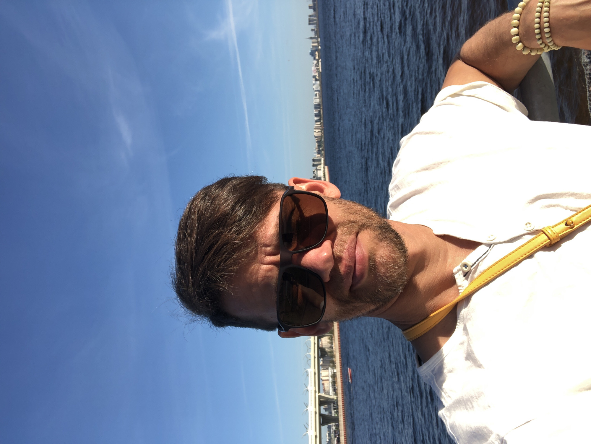 A man wearing sunglasses and a white shirt takes a selfie outdoors. He has short brown hair and a beard, and a yellow strap is visible around his neck. He is standing on a railing overlooking a body of water, with a city skyline and a bridge in the background. The sky is blue with some wispy clouds. This scene is likely located in Saint Petersburg, Russia, near the Gulf of Finland, given the architectural style of the bridge and the general urban landscape visible.