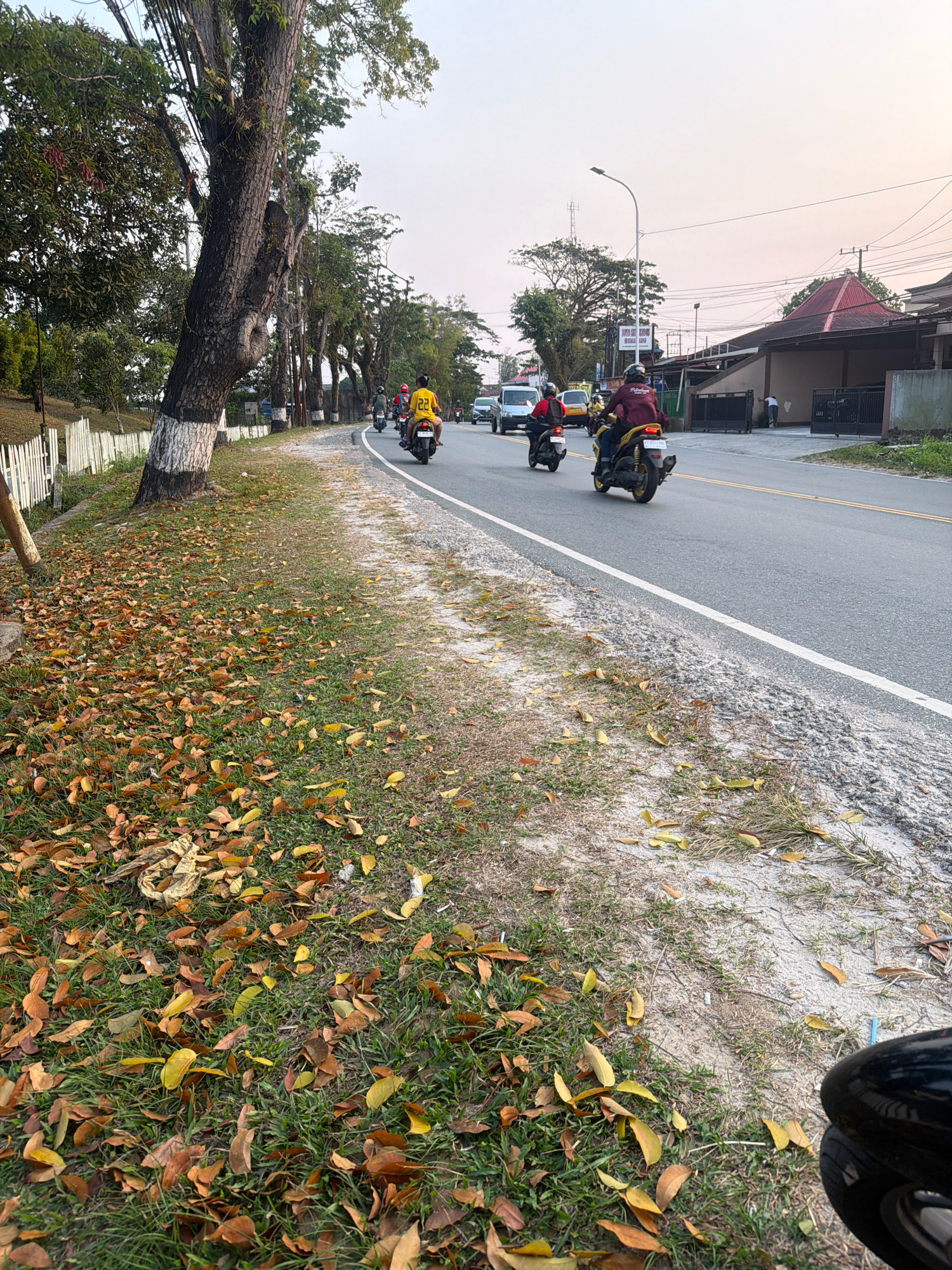 The image shows a street scene in Balikpapan, Indonesia, with several motorcycles riding on the asphalt road. The road is lined with trees and a grassy shoulder covered in fallen leaves. There are buildings and a traffic light visible in the background. A white fence runs along the left side of the road. The sky is hazy with a pale orange hue, suggesting it might be early morning or late afternoon.