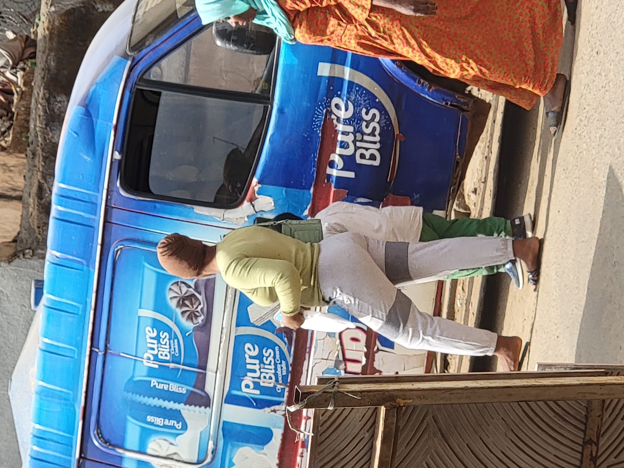 A partially visible blue van with “Pure Bliss” advertising, is parked in the city of Maiburiji, Nigeria. Two people stand near the van. One wears an orange patterned garment and the other is wearing green shirt and light colored pants, holding a piece of paper in their hands. The van features various “Pure Bliss” advertisements, indicating the brand of product being sold.