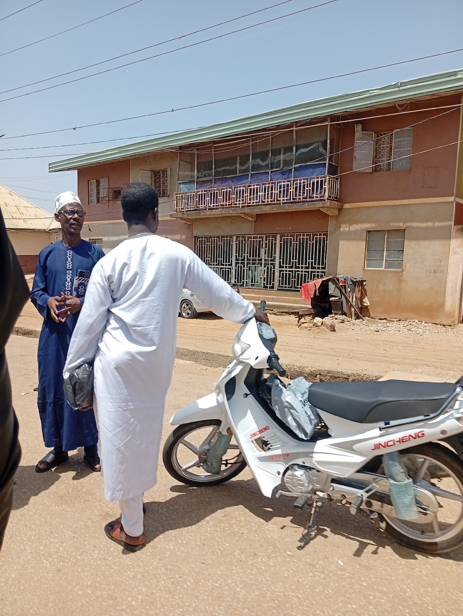 Two men are standing on a dirt road in front of a two-story building in Jos, Nigeria. The man on the left, wearing a blue traditional outfit and a white cap, is speaking to the man on the right, who is wearing a white traditional outfit. A white JinCheng motorcycle is parked to the right of the man in white. The building behind them has balconies with railings and several windows. The road is unpaved and dusty, with some debris scattered around. The sky is clear and blue.