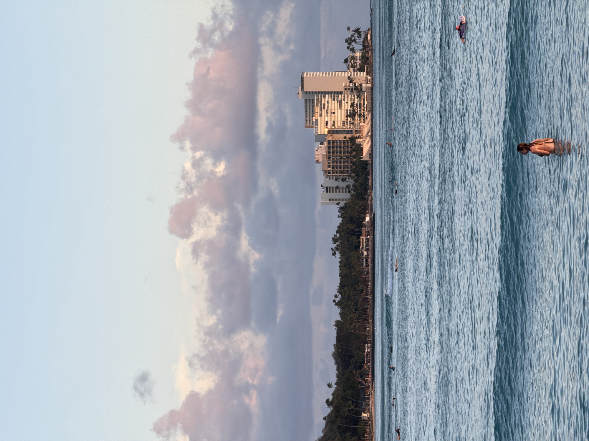 The image depicts a coastal scene in Honolulu, United States of America. The foreground shows a stretch of blue water where several people are swimming and surfing. In the midground, a line of trees runs along the shore, with buildings visible behind them, including some taller structures that suggest a developed urban area. The sky, which occupies a significant portion of the image, is a mix of blue and clouds with hints of pink, indicating either sunrise or sunset. The photograph is presented in a rotated orientation.