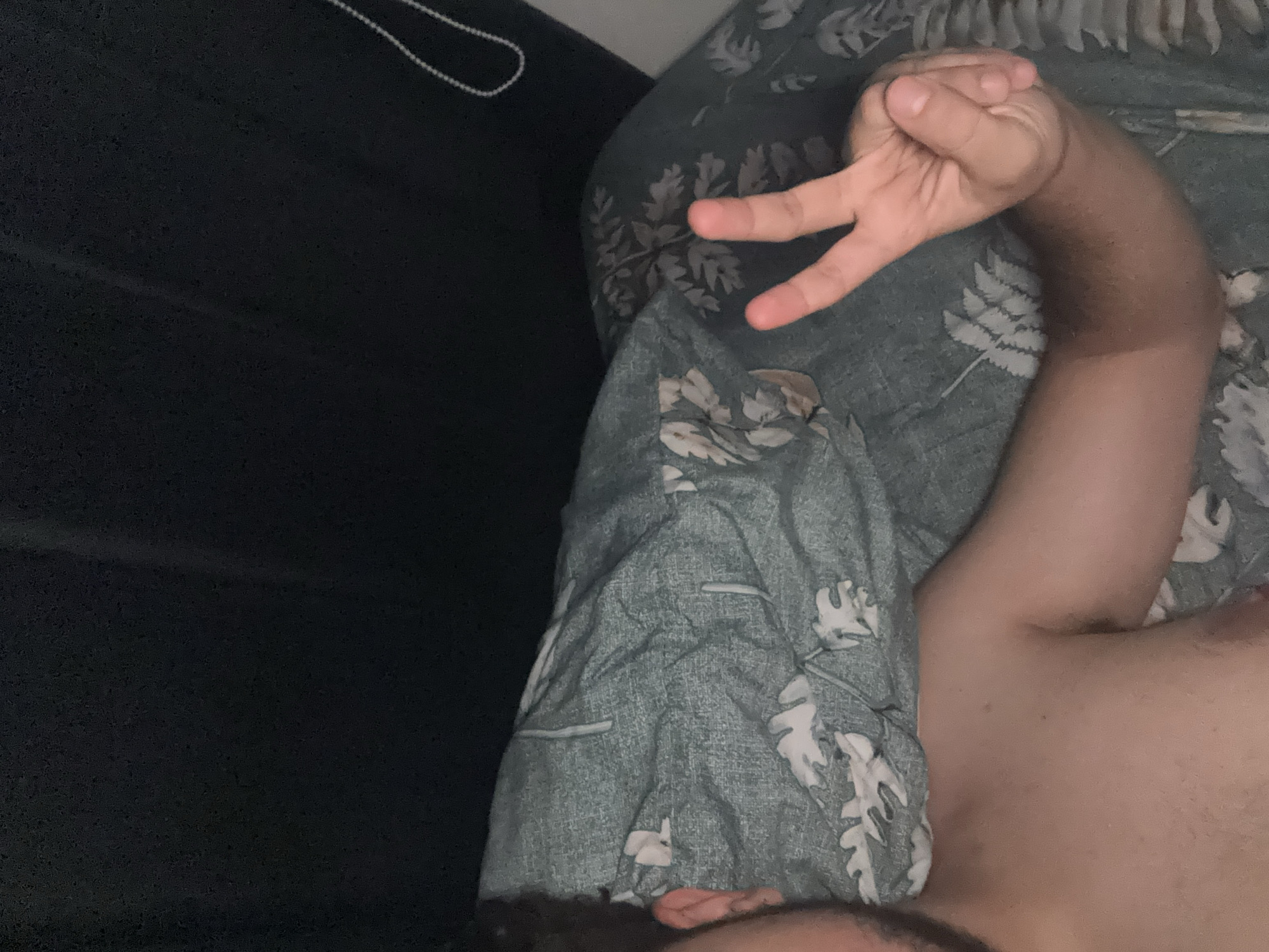 An overhead shot shows a person lying in bed. The person's torso and arm are visible on the left and bottom of the frame, respectively. They are wearing no shirt. The person's right hand is raised, and they are making a peace sign with their index and middle fingers extended. The background consists of a dark, textured wall and a floral patterned comforter. There is a beaded string hanging from the top right corner of the frame. The scene appears to be indoors.