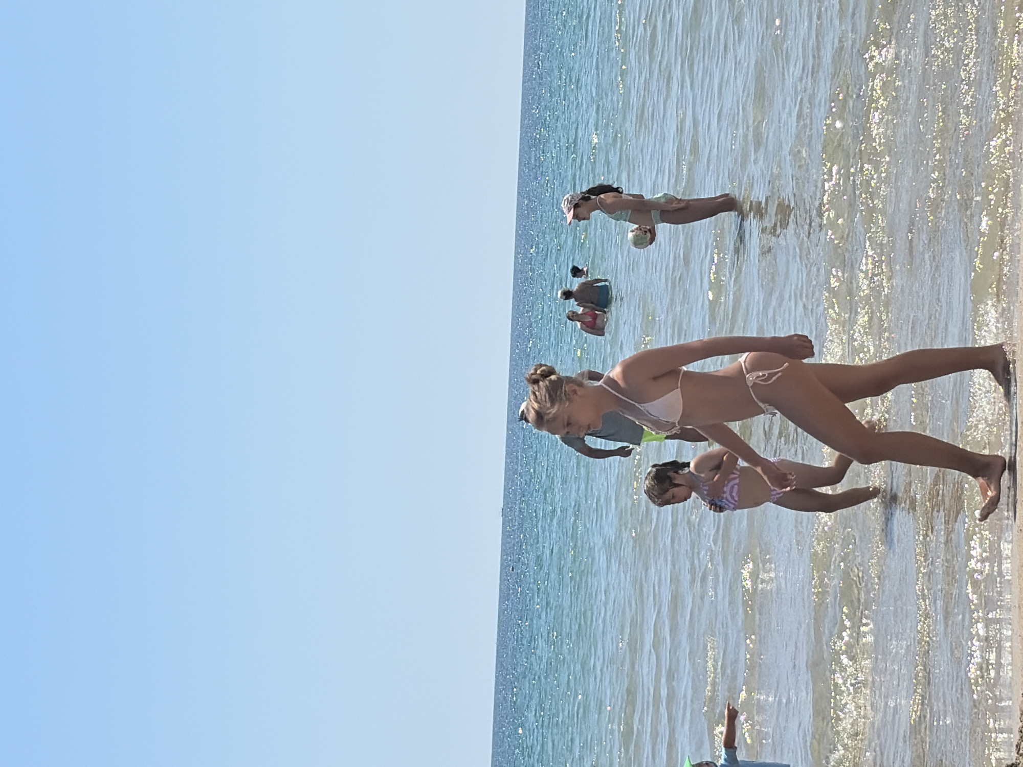 This is a medium shot of people on a sunny day at the beach in Primorsko, Bulgaria. The foreground shows the legs and torso of a young woman in a white bikini walking from right to left on the wet sand. Behind her, a young girl in a purple and white bikini walks from right to left through shallow water. To her left, a person wearing a green hat and a blue shirt points to the right. In the middle ground, several people are standing or wading in the water. To the right, a young girl in a mint green bikini holds a ball. The water is a clear turquoise and reflects the bright sunlight. The sky is a clear, pale blue with no clouds. In the distance, a small boat is visible on the horizon.