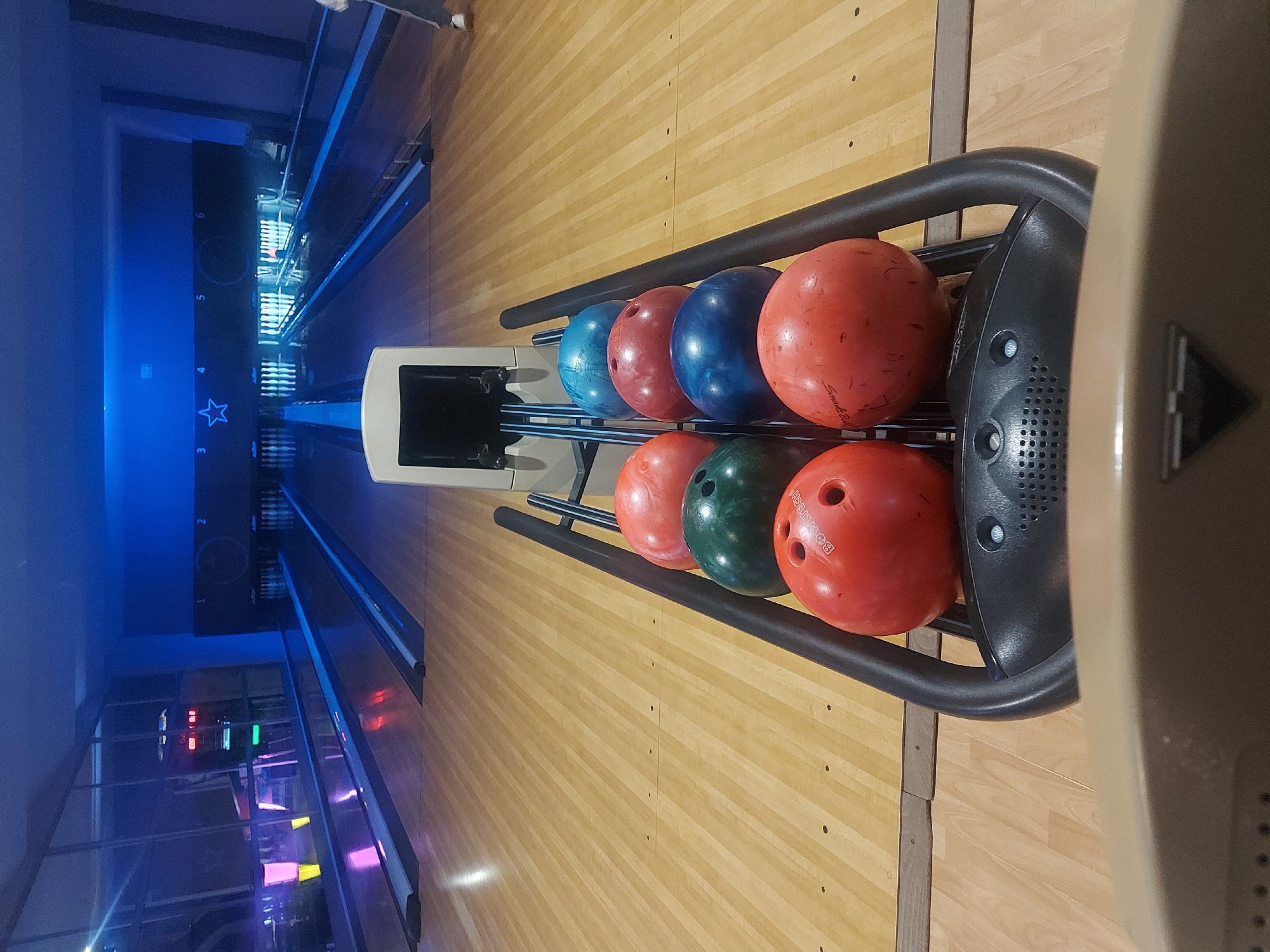 The image shows a bowling alley with bowling balls of various colors (blue, red, green, and orange) neatly arranged on a rack. The alley itself is made of wood and marked with lines. The atmosphere appears to be dimly lit, with blue neon lights illuminating the lanes. The location is not evident from the image alone.