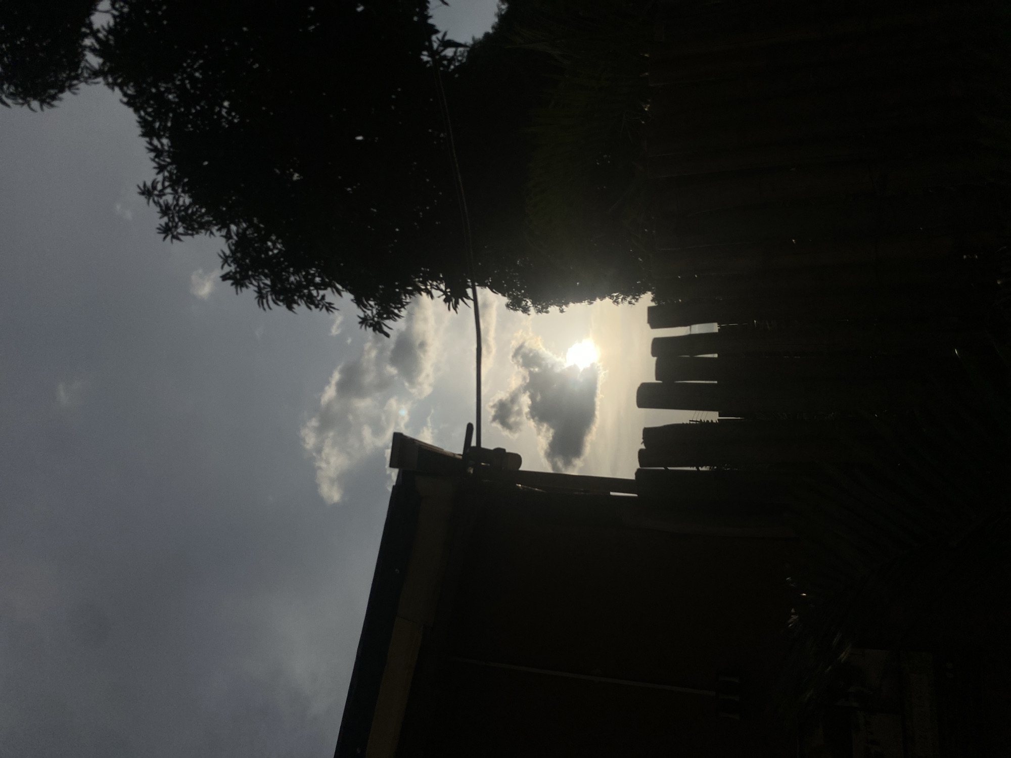 The image captures a silhouetted view of a yard at sunset in Barden Yana, Nigeria. A wooden fence made of vertical poles stands prominently in the foreground, casting deep shadows. To the left, the side of a building with a visible roofline is silhouetted against the sky. A large tree with dark foliage is visible on the right side of the frame, also silhouetted. The sun is setting, creating a bright glow behind scattered clouds, with its rays peeking through the clouds and illuminating the sky. The overall scene is dark due to the low light conditions of sunset, with the silhouettes of the structures and vegetation standing out against the brighter sky.
