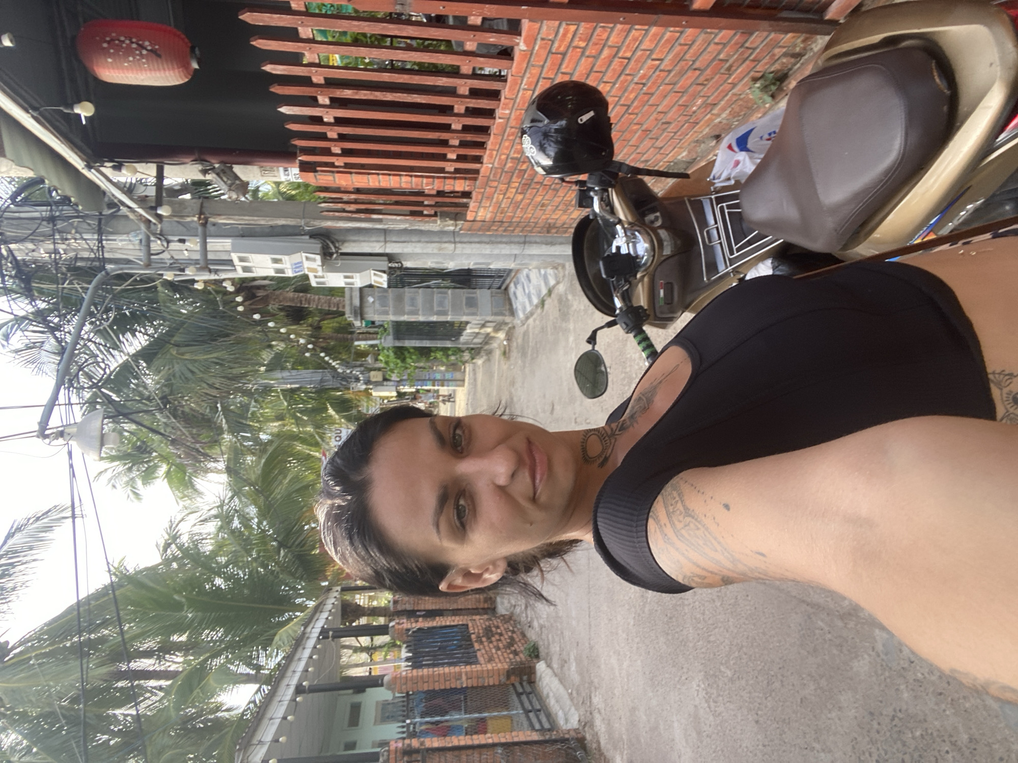 A selfie of a woman with dark hair, green eyes, and visible tattoos on her neck and arm, taken from the chest up. She is wearing a black, sleeveless top and is positioned in front of a scooter with a brown seat and a black helmet on the handlebars. The background suggests a street scene in Mũi Né, Vietnam, characterized by palm trees, brick walls, and a red lantern hanging from a utility pole. The environment appears to be a residential or commercial area with buildings and a paved road.