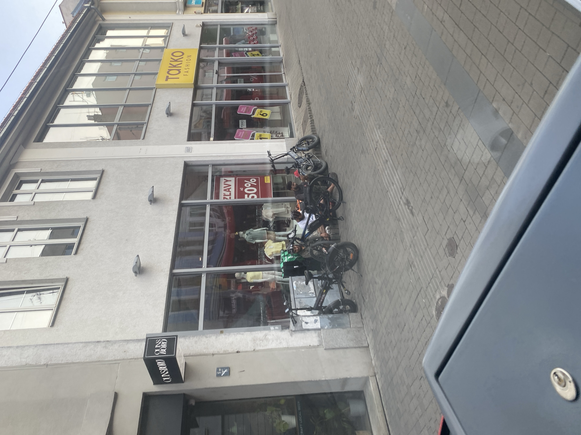 The image shows a street in Bratislava, Slovakia, with storefronts and bicycles. On the left, a sign for "QUNSBORO" is visible above a storefront. Further to the right, a yellow sign with "TAKKO FASHION" is displayed on another building. Several bicycles are parked on the sidewalk in front of the shops. Two individuals are seated near the bicycles. The street is paved with cobblestones, and there are a few potted plants outside one of the shops. The overall scene depicts a commercial area of the city.