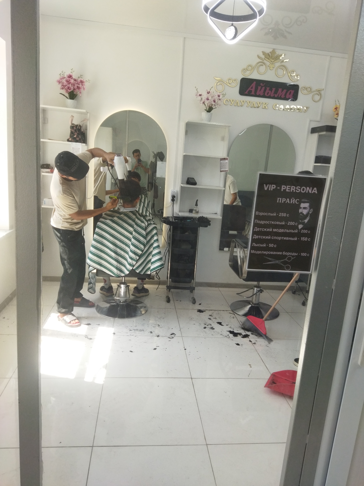 A barber is cutting hair in a salon, using a hairdryer and scissors. The barber, wearing a black baseball cap and tan t-shirt, is standing and facing away from the camera. The client, seated in a barber chair and draped in a green and white striped cape, has their back to the camera. The salon has white tiled floors, with scattered hair clippings visible. A price list for VIP-PERSONA services is displayed on a black board, featuring services such as haircuts for adults, teenagers, and children, as well as beard modeling. In the background, a mirror reflects the salon interior, showing another person in a green shirt holding a phone, possibly taking a photo or recording. The overall scene depicts a typical barbershop environment in Mailuu-Suu City, Kyrgyzstan.