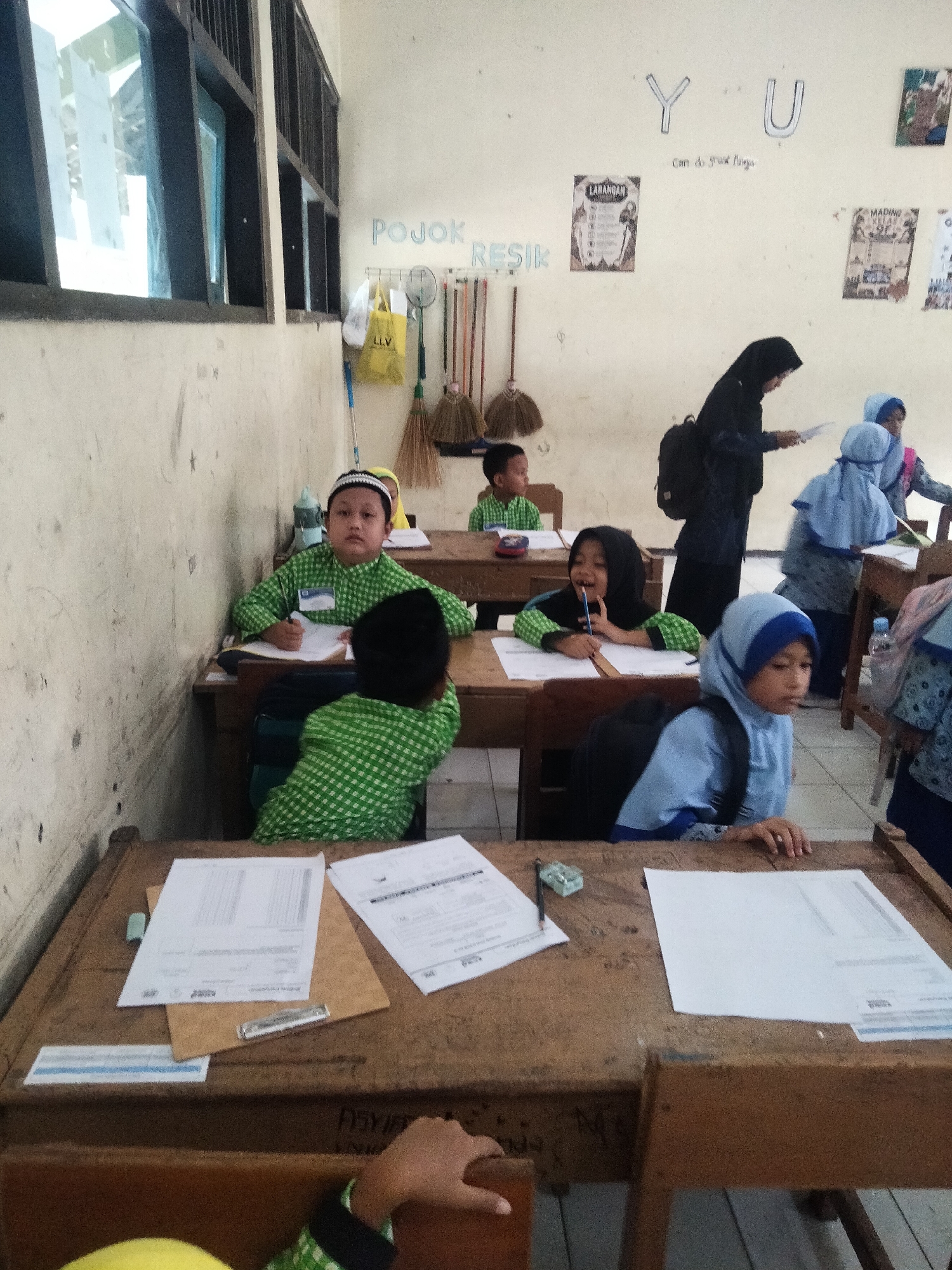 The image depicts a classroom scene likely in Sanggrahan, Indonesia, featuring young students engaged in learning activities. Several children, dressed in traditional Islamic attire including skullcaps and hijabs, are seated at wooden desks. The desks are cluttered with papers and writing utensils, suggesting an ongoing lesson or exam.
In the foreground, a boy in a green checkered shirt and skullcap is focused on his work, holding a pencil. Beside him, another student in a green shirt appears to be writing. Towards the right, a girl wearing a light blue hijab and a similar colored top is looking off to the side, while another student in a darker blue hijab sits behind her.
In the background, an adult, possibly a teacher, is standing and observing the students. The classroom walls are adorned with educational posters and some decorative letters, including "Y U". Various cleaning tools, such as brooms and a mop, are visible near the wall, indicating a tidy learning environment. Sunlight streams in through a large window on the left, illuminating the room. The overall atmosphere appears to be one of quiet concentration and learning.