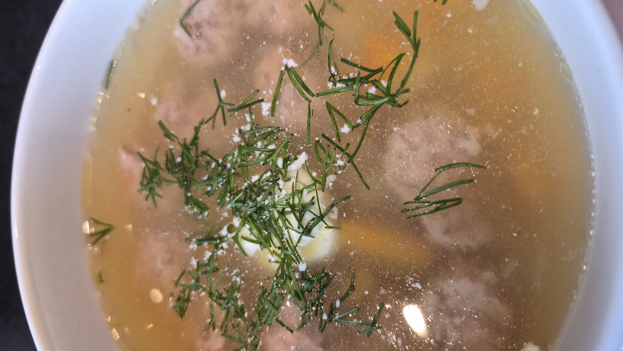 A close-up, overhead shot displays a bowl of light-colored soup, likely a broth-based dish, filled with small, round meatballs and pieces of diced vegetables, possibly carrots. The soup is garnished with finely chopped fresh dill, which is scattered across the surface. A small dollop of a light-colored, creamy substance, perhaps sour cream or yogurt, is visible beneath the dill. The lighting suggests an indoor setting, possibly a kitchen or dining area. The image does not contain any identifiable landmarks or elements that would associate it with Daugavpils, Latvia.