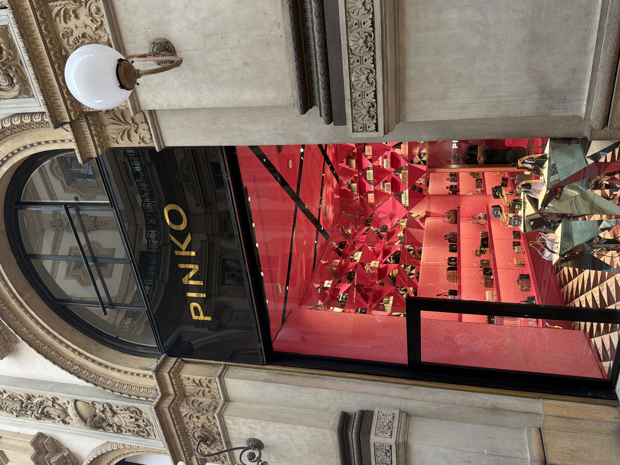 The image shows the entrance to a "PINKO" store, likely in Milan, Italy. The storefront has an arched window above a black-framed doorway with the store's name in gold lettering. The interior of the store is visible through the doorway, featuring a display of red angular decorations and shelves stocked with handbags. The exterior architecture is ornate, with intricate carvings and a white globe light fixture. The perspective is tilted, suggesting the photo was taken from a slightly angled position.