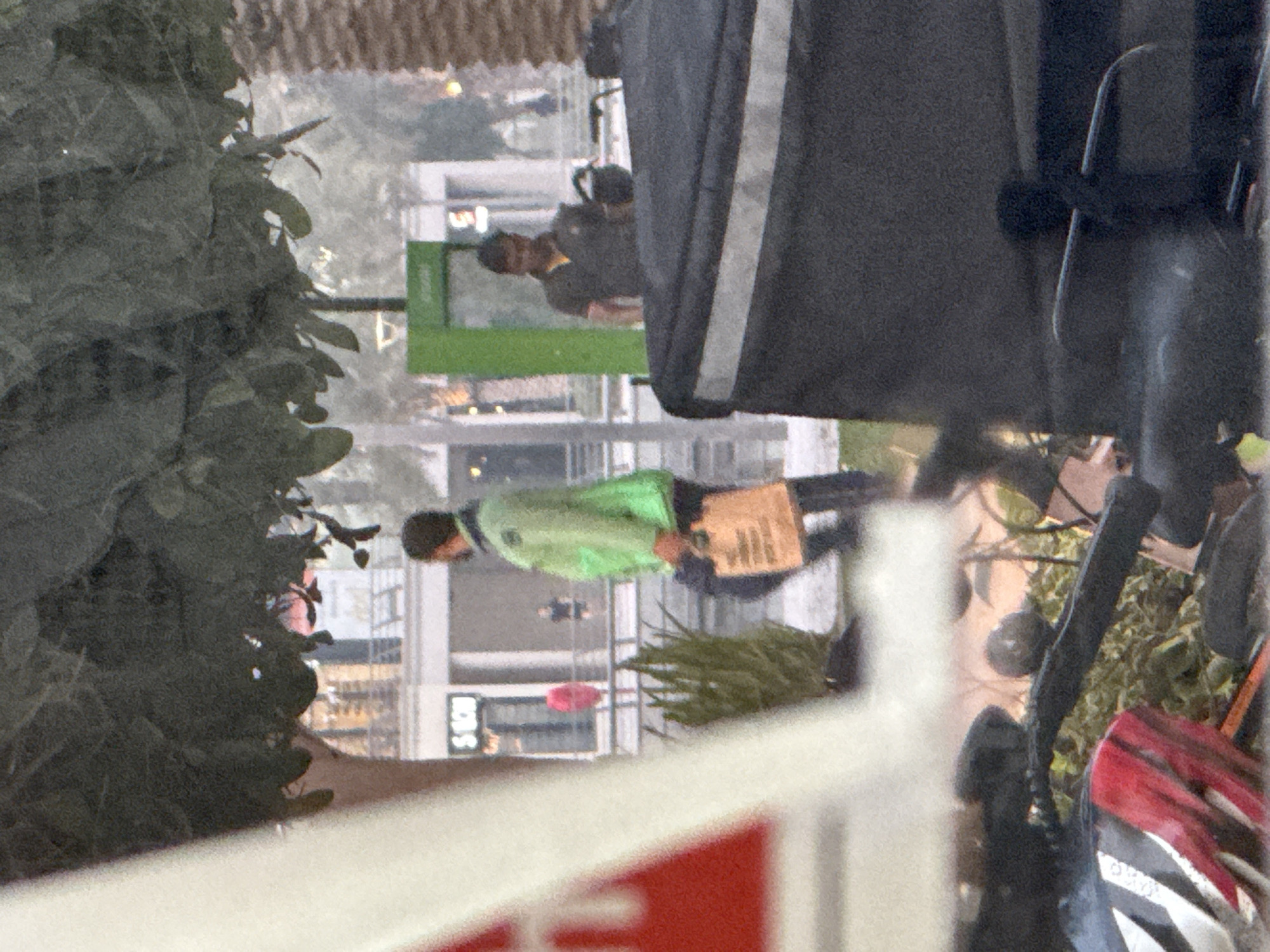 The image shows two men outdoors, possibly in a commercial or pedestrian area in Dubai, United Arab Emirates. In the foreground, partially obscured by a large, out-of-focus object that appears to be part of a vehicle, a man in a bright green jacket and dark pants is walking away from the camera. He is carrying a light brown paper bag in his right hand. In the background, another man is standing near a green structure, wearing a dark polo shirt with a yellow collar and dark pants. He is looking towards the left of the frame. The environment appears to be urban, with buildings and palm trees visible in the distance. The lighting suggests it is daytime. The overall scene conveys a sense of everyday activity in a public space.