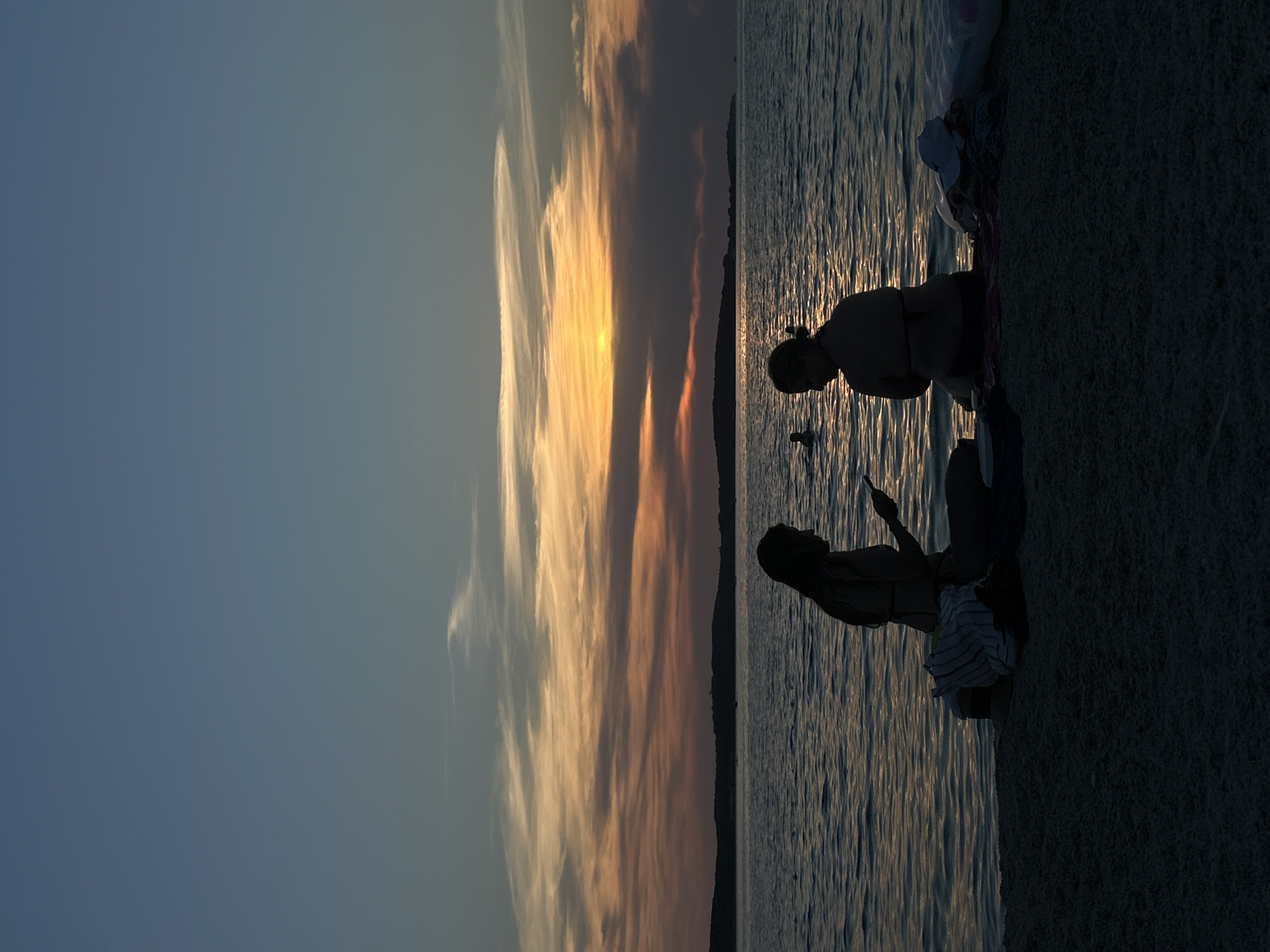 Two silhouetted figures, appearing to be women, sit on a sandy beach facing the sea at dusk. The figure on the left, in profile, is looking down at a mobile phone in her hands. The figure on the right, also in profile, is looking towards the sea. There are several items of clothing and what appears to be a cap or hat piled beside the figures on the sand. The sea is calm with gentle ripples reflecting the warm colors of the sunset. The sky displays a gradient of blues and oranges, with thin clouds streaked across the horizon where the sun is setting. A silhouetted landmass is visible in the distance. The scene suggests a peaceful end to a day at the beach.