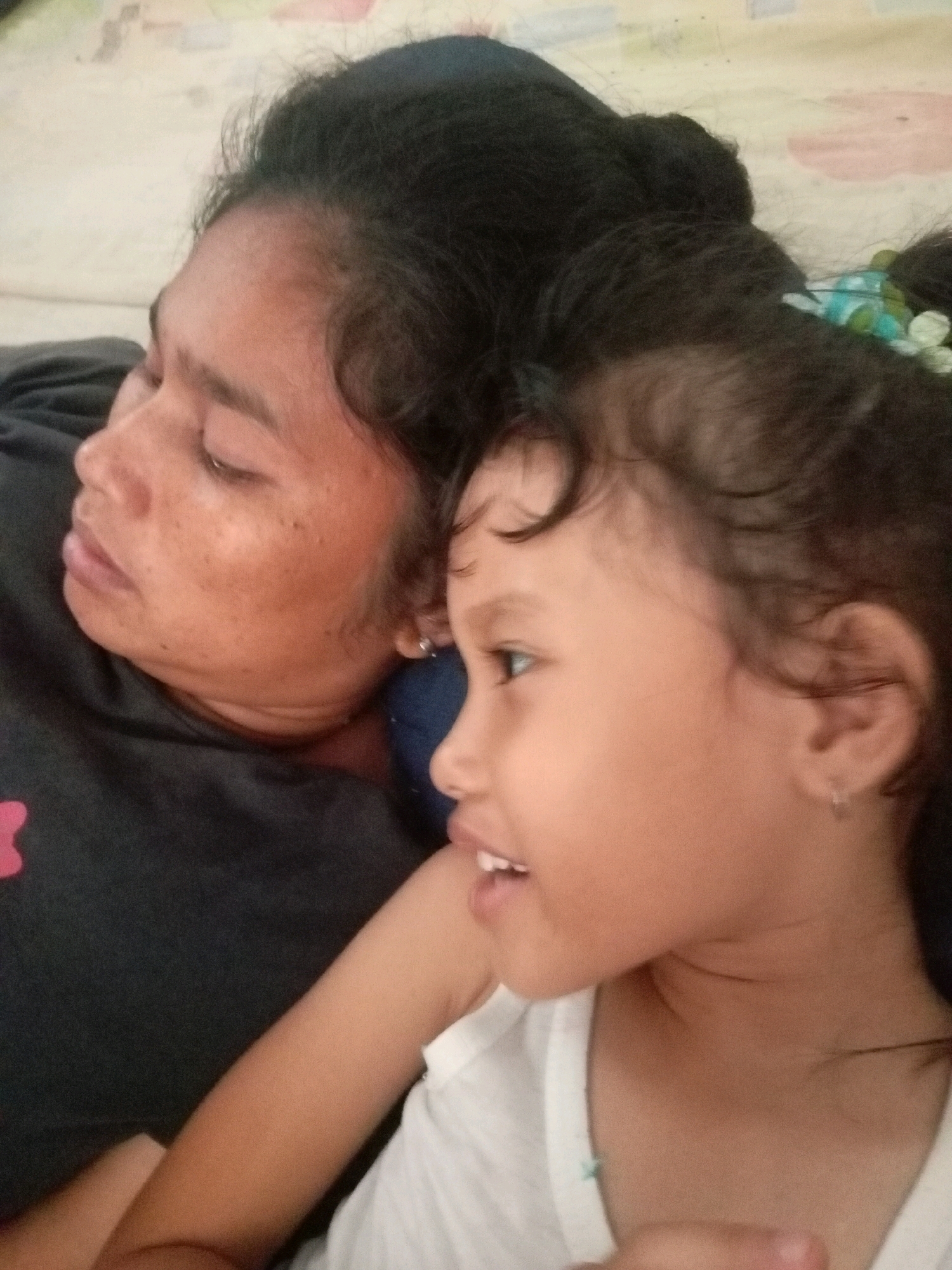 An adult female and a child are lying down, facing the camera from a side profile. The adult, on the left, has dark hair styled in a bun. Her eyes are closed, and her head is tilted slightly back. She is wearing a dark grey t-shirt with a pink emblem on the left side of her chest. The child, on the right, has dark hair with some curls around her face, adorned with a light blue and green hair accessory. The child is looking towards the left with an open mouth, revealing teeth. She is wearing a white t-shirt. Both individuals appear to be indoors, resting on a surface that resembles a bed or mattress with a patterned fabric. The lighting suggests an indoor setting, possibly a bedroom. The image appears to be a personal photograph taken in Batam, Indonesia.