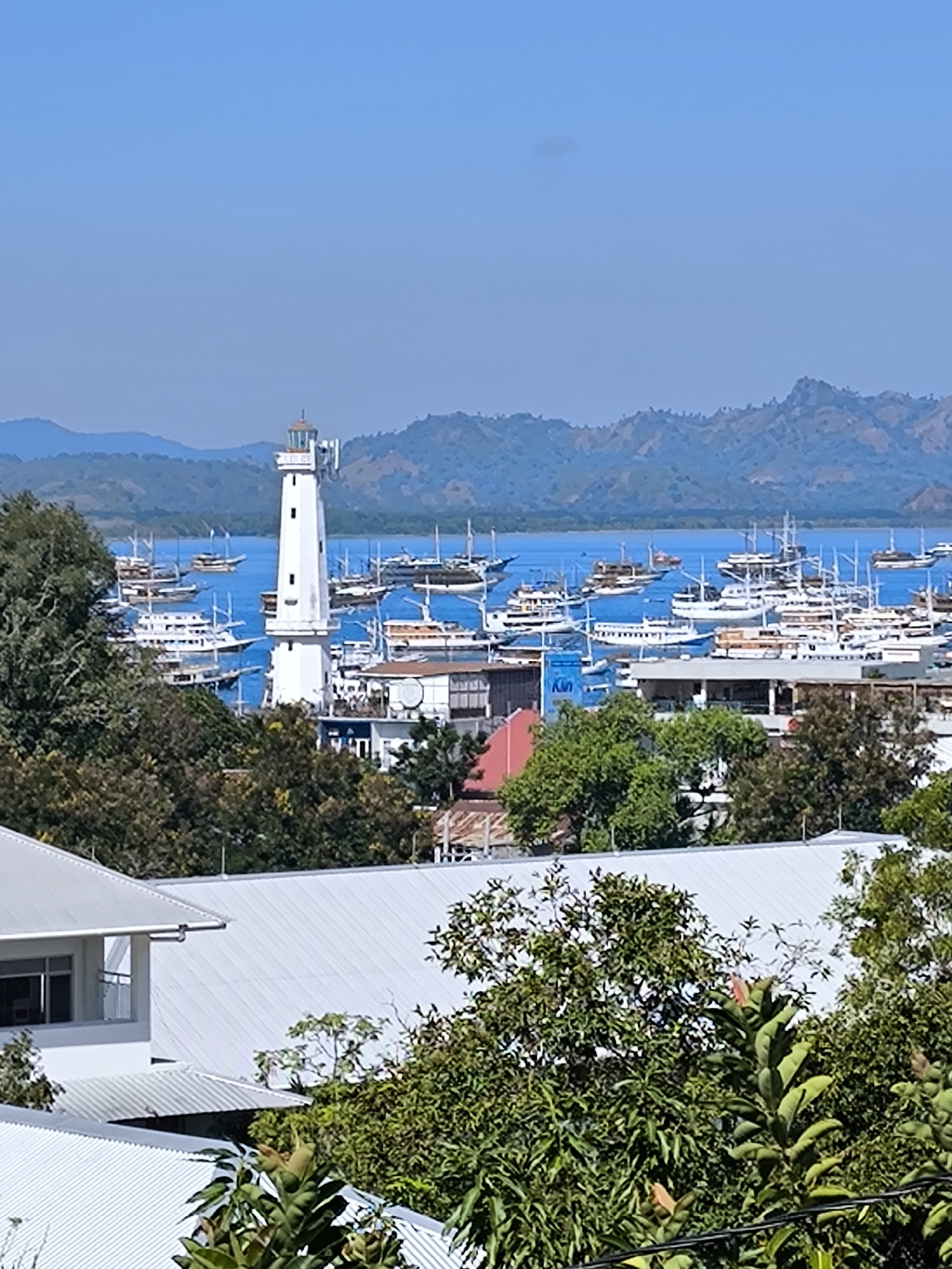 The image depicts a scenic view of Labuan Bajo, Indonesia, with a prominent white lighthouse standing tall on the right side of the frame. The lighthouse is surrounded by a bustling harbor filled with numerous boats of various sizes, from small fishing vessels to larger sailboats. The boats are docked in clear blue waters, with a backdrop of lush green hills and mountains in the distance. The sky is a vibrant blue, with a few scattered clouds. In the foreground, the rooftops of white buildings and dense foliage are visible, suggesting a coastal town or city. The scene conveys a sense of tranquility and natural beauty, with the lighthouse serving as a symbol of the maritime heritage of the region.