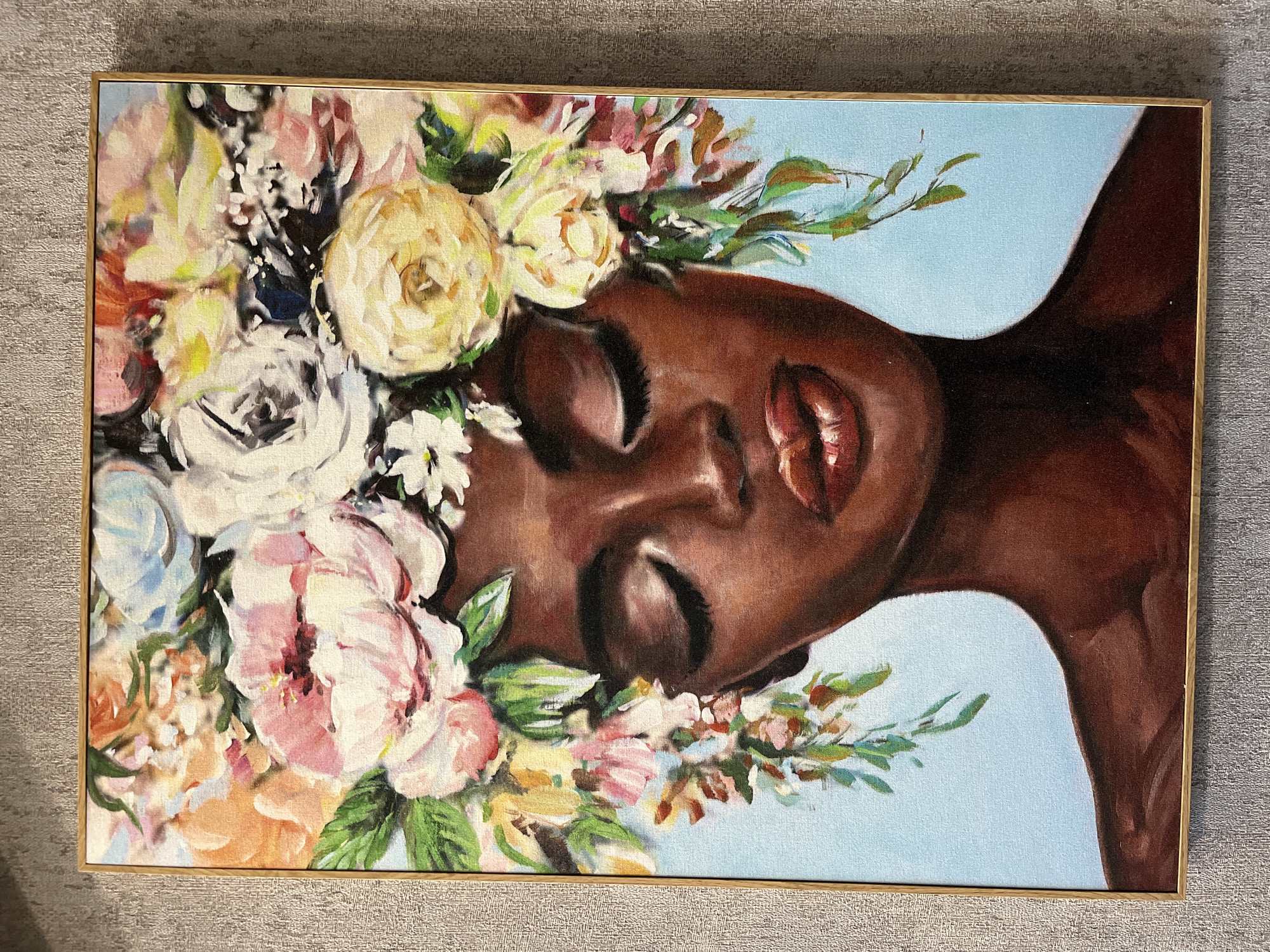 A framed painting is prominently displayed against a textured gray wall. The artwork depicts a serene portrait of a woman with rich, dark skin, her head gently tilted. Her eyes are closed, and her lips are slightly parted, conveying an expression of peace, rest, or profound contemplation.
An abundant and vibrant floral arrangement encircles her head, forming what appears to be a lush flower crown or an intricate hair adornment. These flowers are rendered in a painterly, impressionistic style, showcasing a variety of blooms in soft hues, including cream, white, light pink, peach, and hints of light blue, interspersed with green leaves. Some prominent flowers resemble roses or peonies. The background of the portrait is a solid, tranquil light blue, providing a soft contrast to the subject's skin tone and the bright blossoms.
The painting is enclosed within a thin, light-colored frame, likely made of natural wood or finished in a subtle gold tone. The overall mood of the artwork is one of beauty, tranquility, and natural elegance. There is no visible text within the painting. The image itself does not provide specific location cues, weather conditions, or a precise time of day beyond the serene atmosphere created by the art.