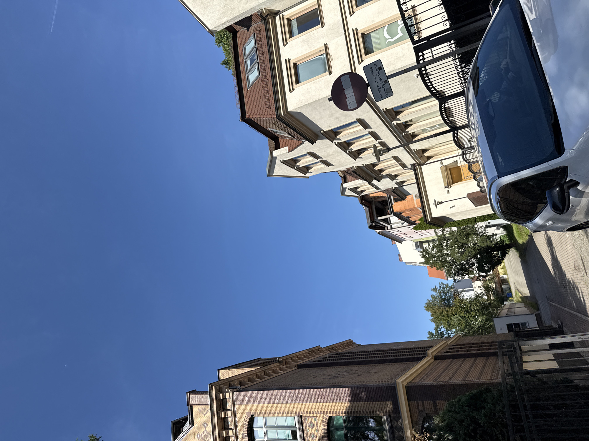 The image depicts a street scene in Zgorzelec, Poland, on a bright, clear day. A silver SUV is parked in the foreground, angled towards the viewer. The street is paved with light-colored cobblestones and curves uphill, lined with residential buildings.
To the left, a multi-story building with a brick facade in shades of yellow and brown features arched windows. Further down the street on the right, a cream-colored building with a brown tiled roof and dormer windows is visible. A black metal fence with decorative elements runs along the sidewalk in front of this building.
A road sign with a red circle and a horizontal bar, indicating "No stopping" or a similar restriction, is mounted on a pole. Below it, a white sign with black text reads "Nie dotyczy pracowników Liceum Ogólnokształcącego", which translates to "Does not apply to employees of the High School". Trees with green foliage are interspersed among the buildings, and the sky is a vibrant, cloudless blue. The angle of the shot is upward, emphasizing the buildings and the sky.