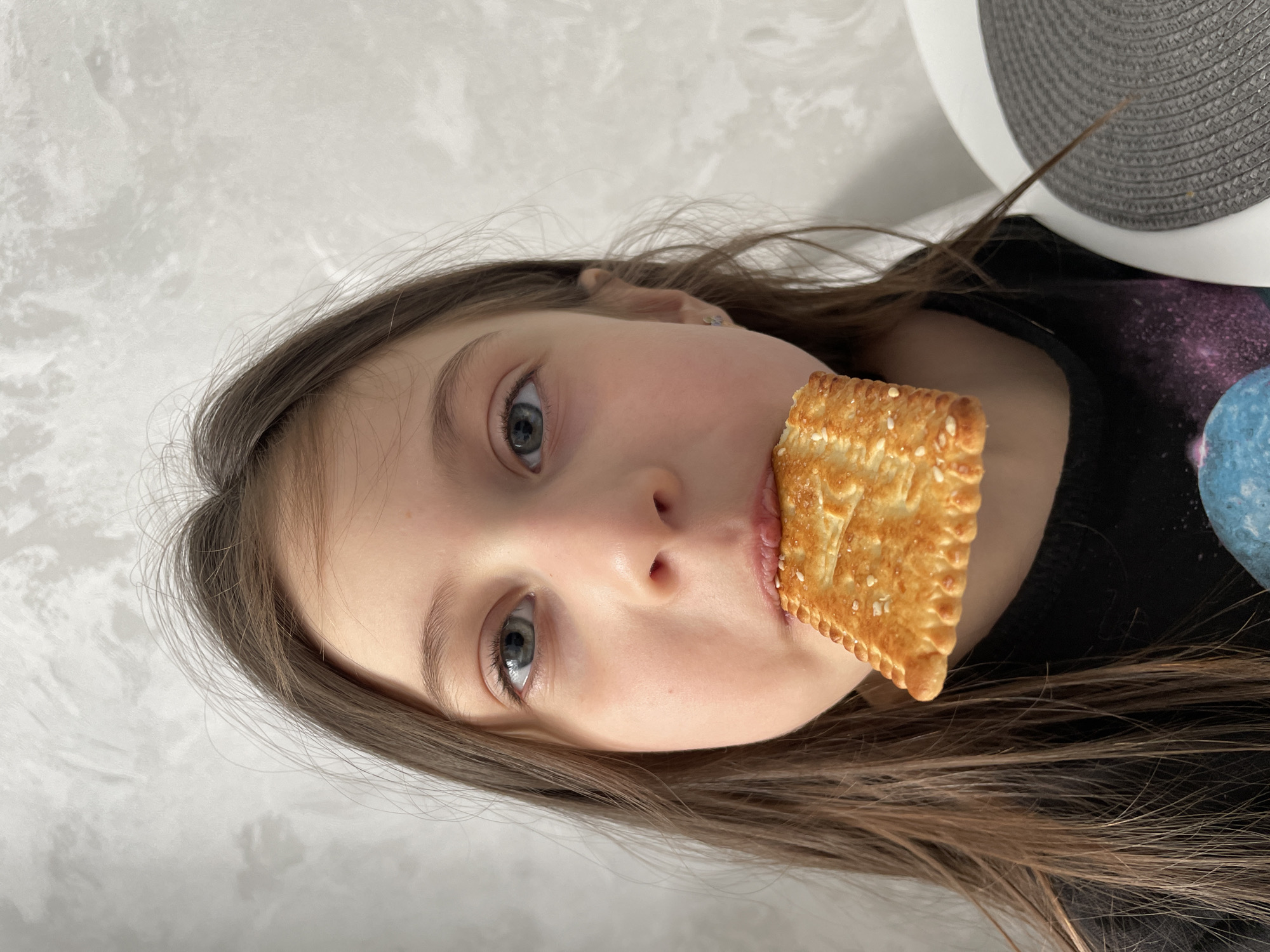 A young girl with long brown hair holds a square cracker in her mouth, facing the camera directly. Her eyes are blue. She is wearing a black shirt with a galaxy pattern. The background is a textured white wall. A circular, gray woven object with a white edge is visible behind her head. There is no indication of location or time.