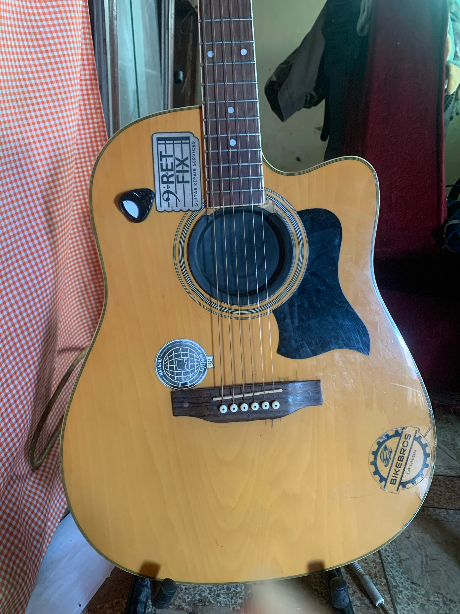 An acoustic guitar with a light-colored wood finish is the central subject of the image. It is leaning against an orange and white checkered fabric. The guitar has several stickers on its body: one is a black and white sticker with "9-RET-FIX GUITAR REPAIR SERVICES" printed on it, and another is a round, silver sticker with a globe graphic and "MALACHI SHREDHOUSE" and "2004" printed on it. A third sticker, a circular blue and yellow sticker with a gear design and "BIKEBROS LA UNION" printed on it, is visible on the lower right side of the guitar's body. The guitar's strings and bridge are visible, as is the soundhole with a black insert. The background is blurred but appears to include a dark red fabric and possibly clothing hanging. The image is taken from a medium distance, focusing on the body of the guitar. There is no indication of the city of San Fernando, Philippines, within the image itself.
