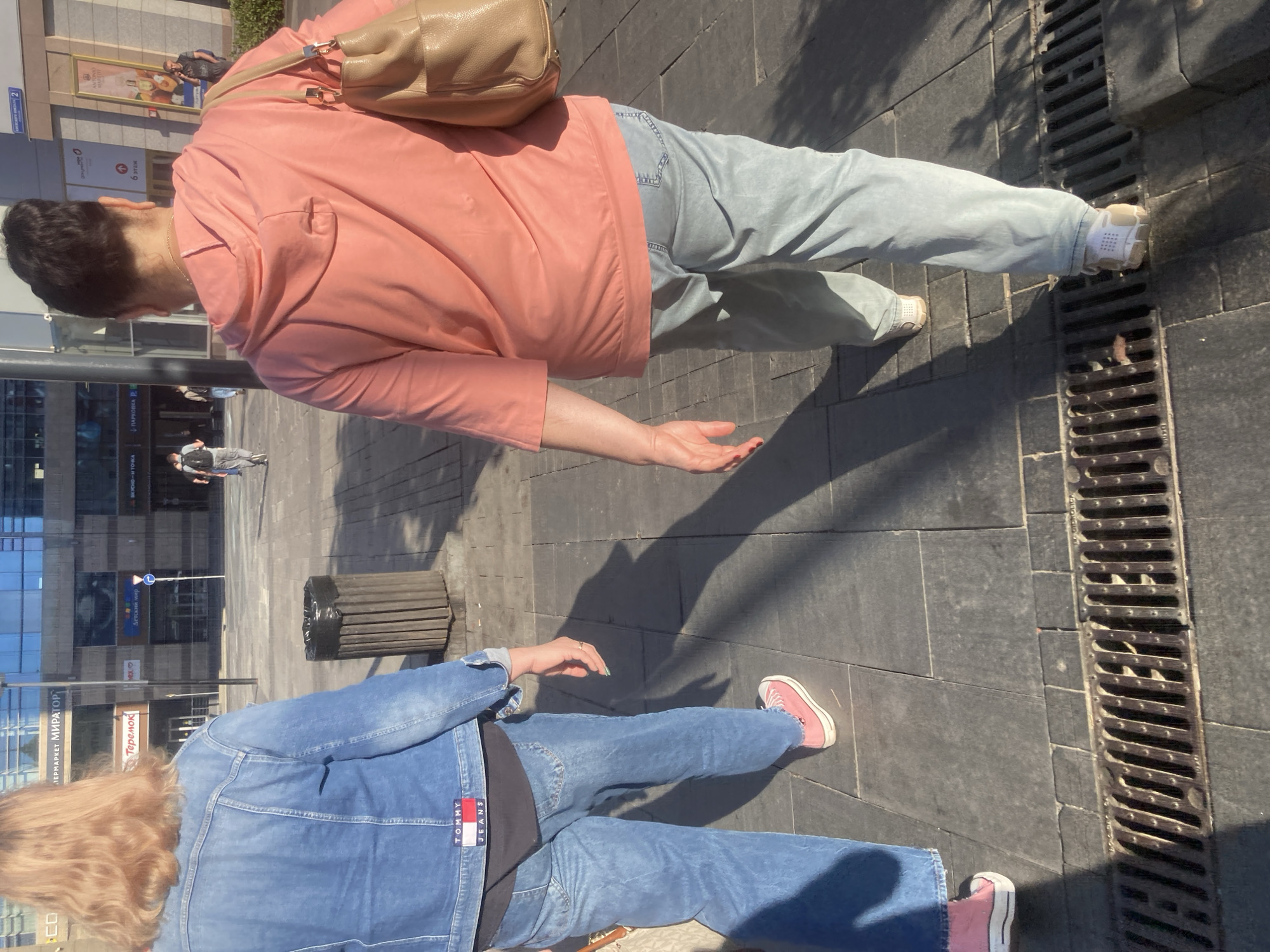 The image shows two people standing on a paved area in Moscow, Russia. One person is wearing a peach-colored jacket, light blue jeans, and a beige shoulder bag. The second person is wearing a blue denim jacket and jeans, with a glimpse of a black shirt underneath. Both individuals are wearing sneakers. A metal grate runs along the side of the pavement. In the background, there is a building with signs indicating "ПЕРМАРКЕТ МИРАТОРГ", "Теремок", "Детский мир", and "ПАРКОВКА".