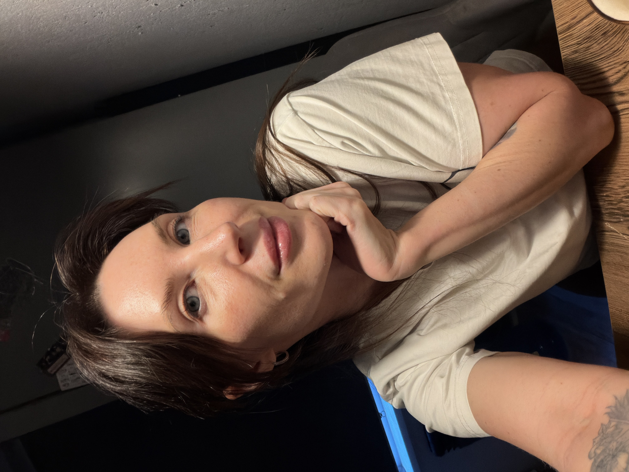 A woman with dark hair and fair skin poses for a selfie. She is wearing a white t-shirt. Her left hand is near her face, supporting her head. She is indoors. The location is unknown and cannot be determined from the image.