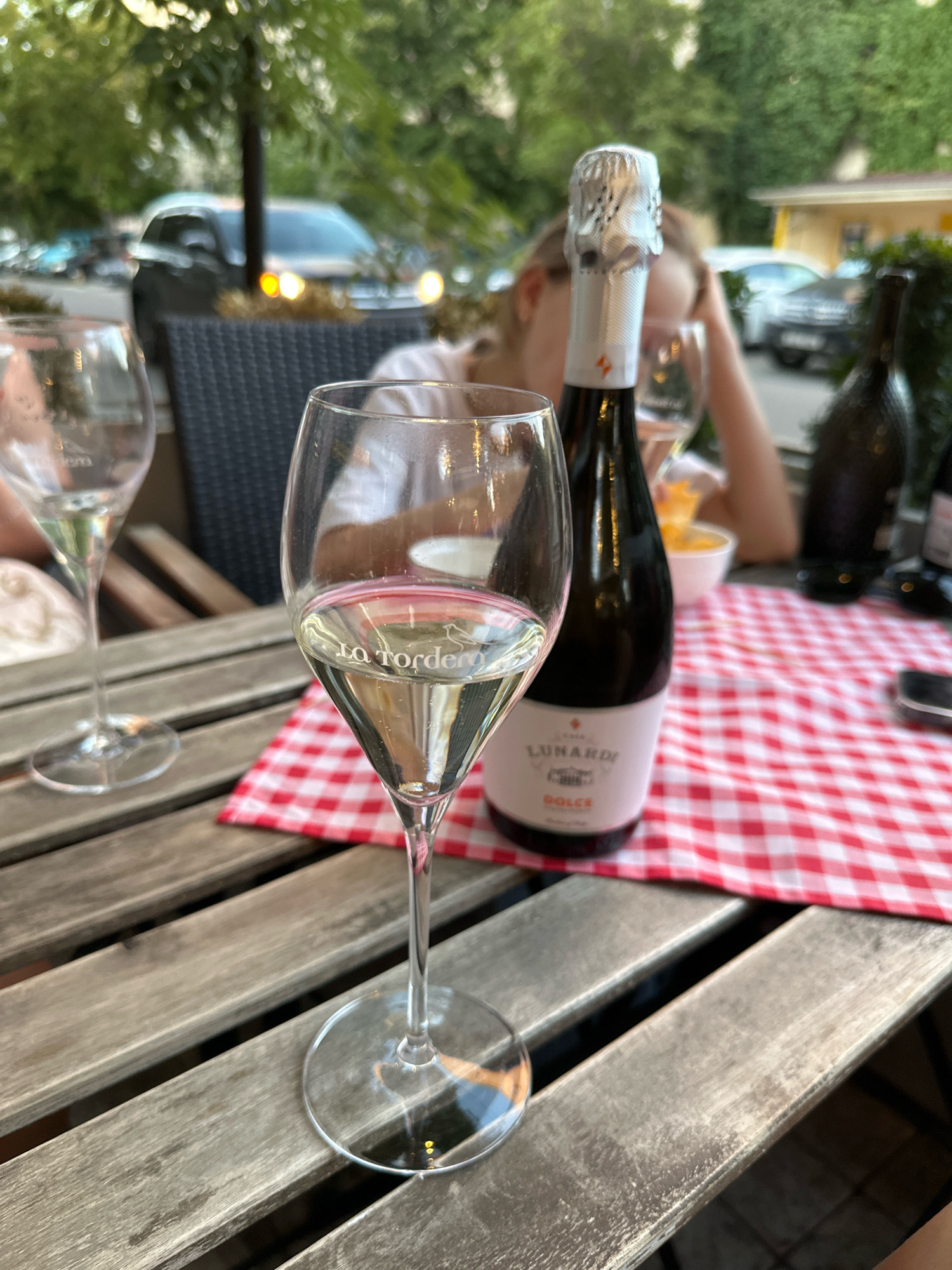 A close-up, eye-level shot captures two wine glasses and a bottle of sparkling wine on a wooden table. The table is set with a red and white checkered tablecloth. The glass in the foreground, partially filled with white wine, has the words "la Tordera" etched into it. Behind it, an empty wine glass stands. To the right, a bottle of "Lunardi Dolce" sparkling wine is prominently displayed, its foil-covered top catching the light. In the background, out of focus, a person is visible, and beyond them, cars and trees suggest an outdoor setting in a city. The overall lighting is natural, indicating daytime. The scene appears to be set at a restaurant or cafe in Tiraspol, Moldova.
