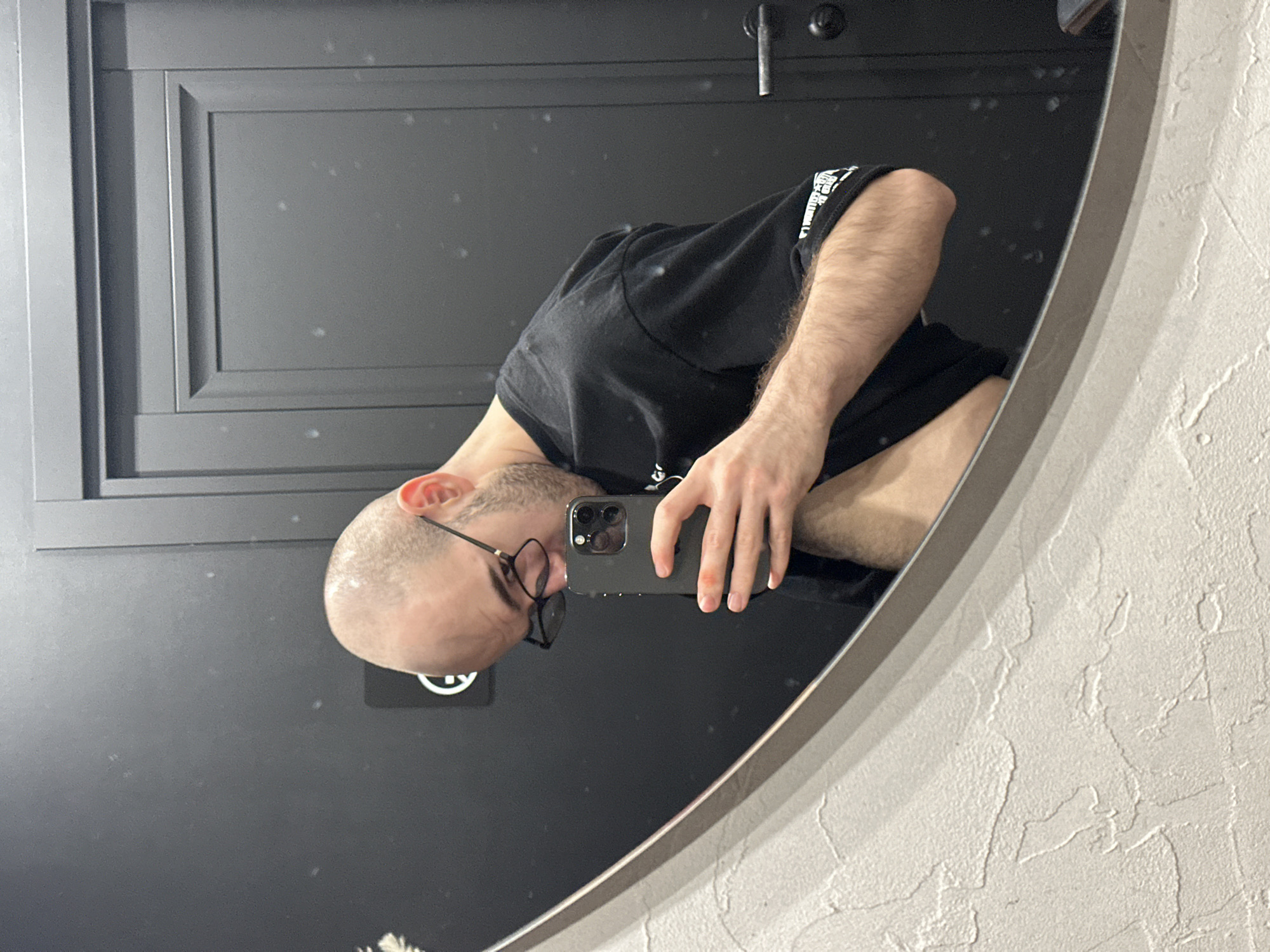 A bald man in a black t-shirt takes a selfie in a mirror. He is wearing glasses and holding a black smartphone. The mirror is hanging on a wall with a dark grey, panelled door nearby and a textured white wall to the side. The location is unclear. There is no discernible evidence of Moscow, Russia.