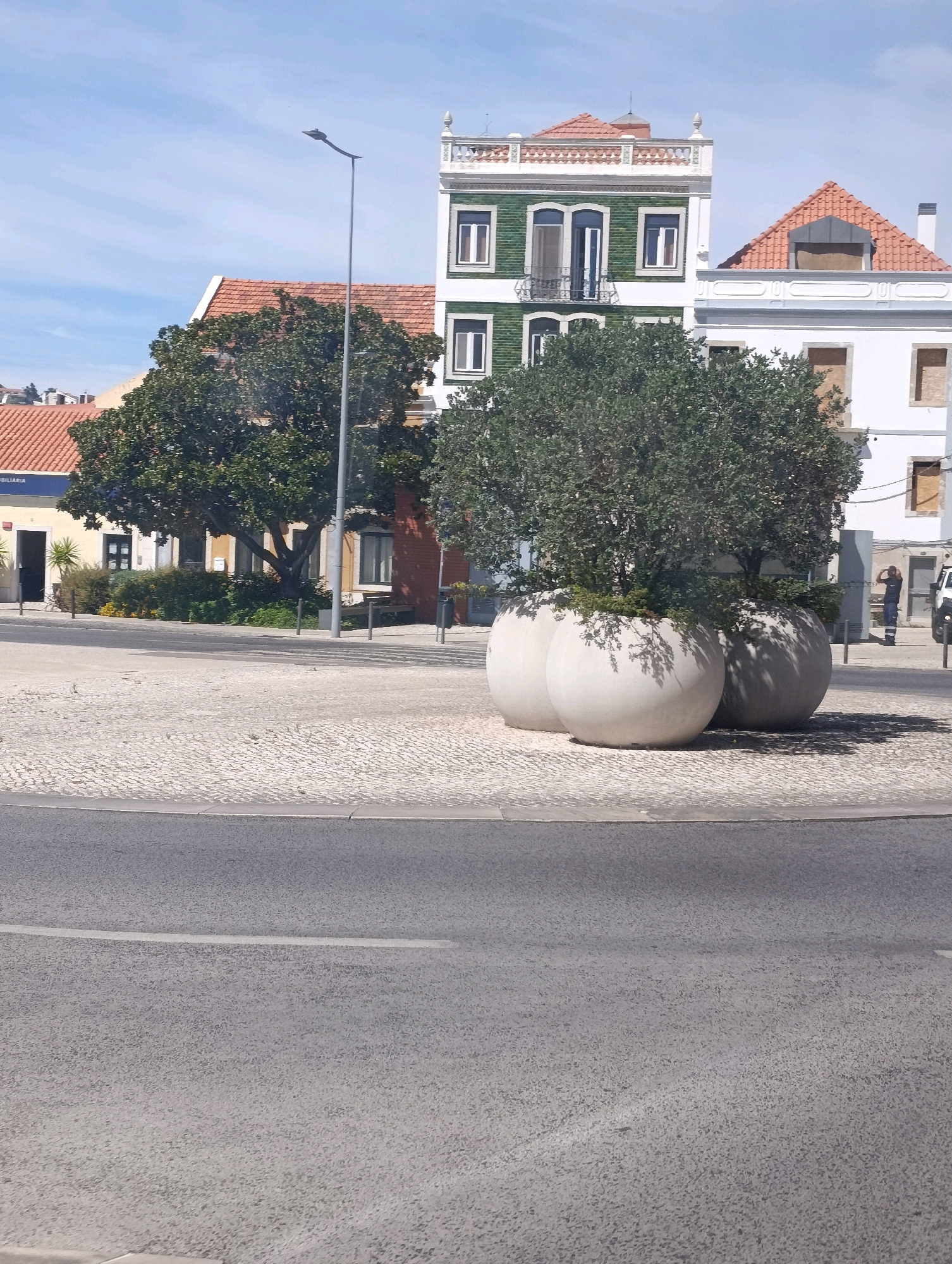 The image depicts a street scene in Setúbal, Portugal. In the foreground, a paved road with a white line runs diagonally across the frame. Behind the road, a cobbled pedestrian area is visible, featuring large, spherical planters containing trees. Several buildings with tiled roofs and varying architectural styles are present in the background, some appearing to be under renovation with boarded-up windows. A lamppost stands on the left side of the cobbled area. In the middle ground, a few people are visible, including one individual standing near a building entrance and another walking away from the camera. The sky is blue with scattered clouds.