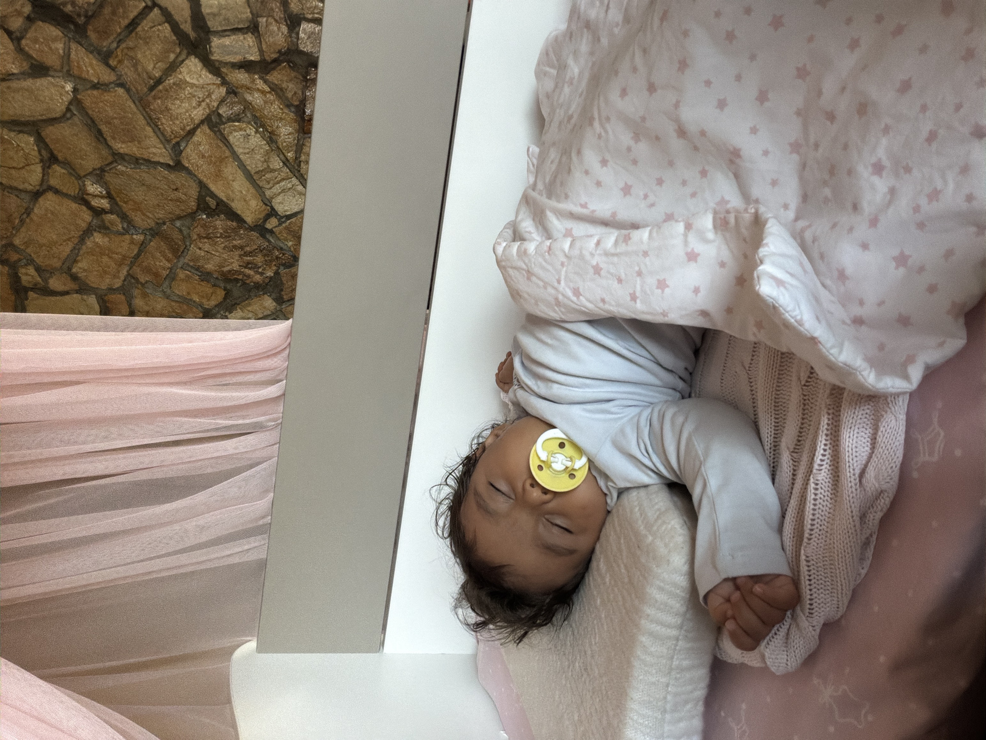 A baby is sleeping peacefully in a crib. The baby is lying on its side with a pacifier in its mouth. The crib is covered with a pink and white star-patterned blanket. The background of the crib includes a white headboard and a wall covered in stone. A pink sheer curtain is visible to the left of the crib. The scene appears to be in a bedroom.