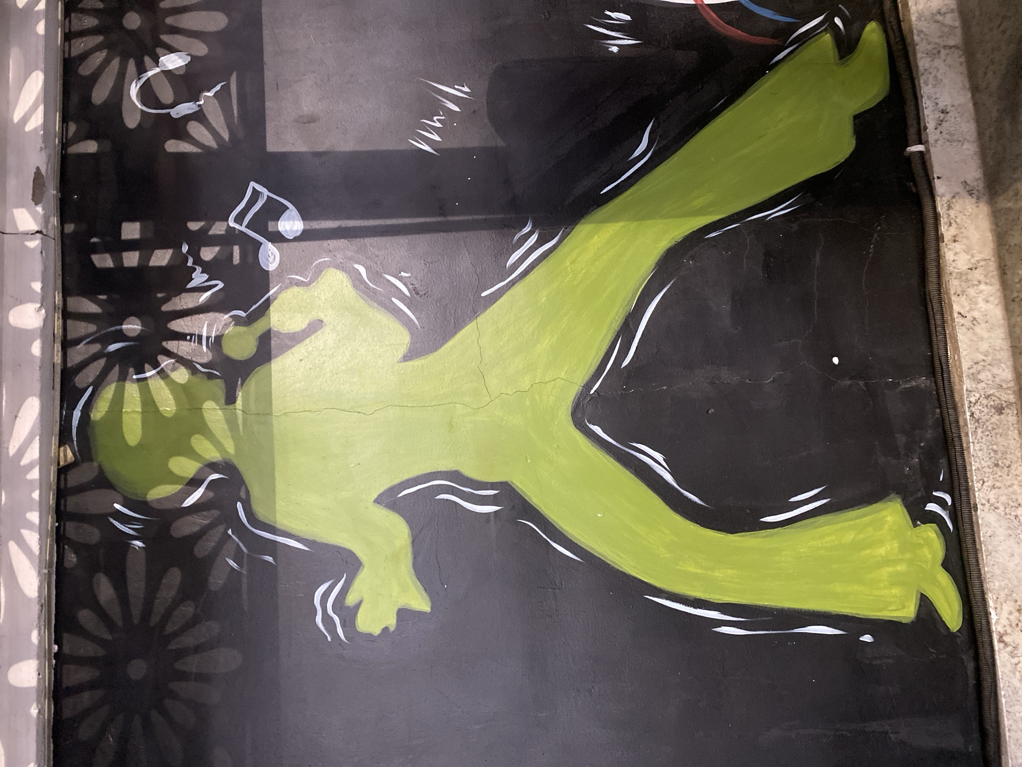 A close-up shot depicts a piece of artwork. The artwork features a stylized, lime-green silhouette of a person lying down on a black background. The person's arms and legs are extended, and white lines accent the outline of the figure, suggesting movement or energy. To the left of the figure's head, there are painted musical notes in white. Part of the background features a pattern of stylized, repeating flowers. The artwork appears to be painted on a rough surface with cracks visible. The location and city are not visible within the image.