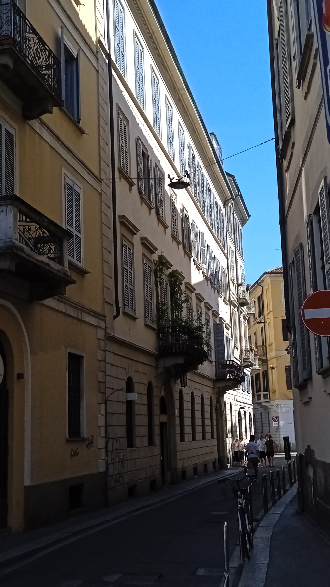 The image shows a narrow street in Milan, Italy, with buildings on both sides. The buildings are tall and have multiple floors with windows and balconies. The street is paved and there are people walking in the distance. The sky is blue and clear, indicating a sunny day. A red and white traffic sign is visible on the right side of the street. The architecture suggests an urban environment with a mix of residential and commercial spaces. The lighting indicates it is daytime, with shadows cast on the street and buildings. The scene appears to be in a historic district, given the style of the buildings.