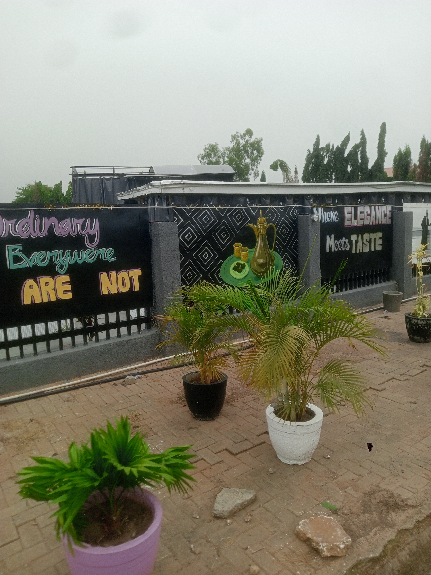 The image shows an outdoor area in Gwarinpa, Nigeria. A black fence with decorative elements and painted signs reading "Ordinary Everywhere ARE NOT" and "Where ELEGANCE Meets TASTE" is prominent. Illustrations of a gold coffee pot and cups on a green plate are also painted on the fence. Several potted palm plants are arranged in front of the fence. The ground is paved with red bricks. The sky is overcast.