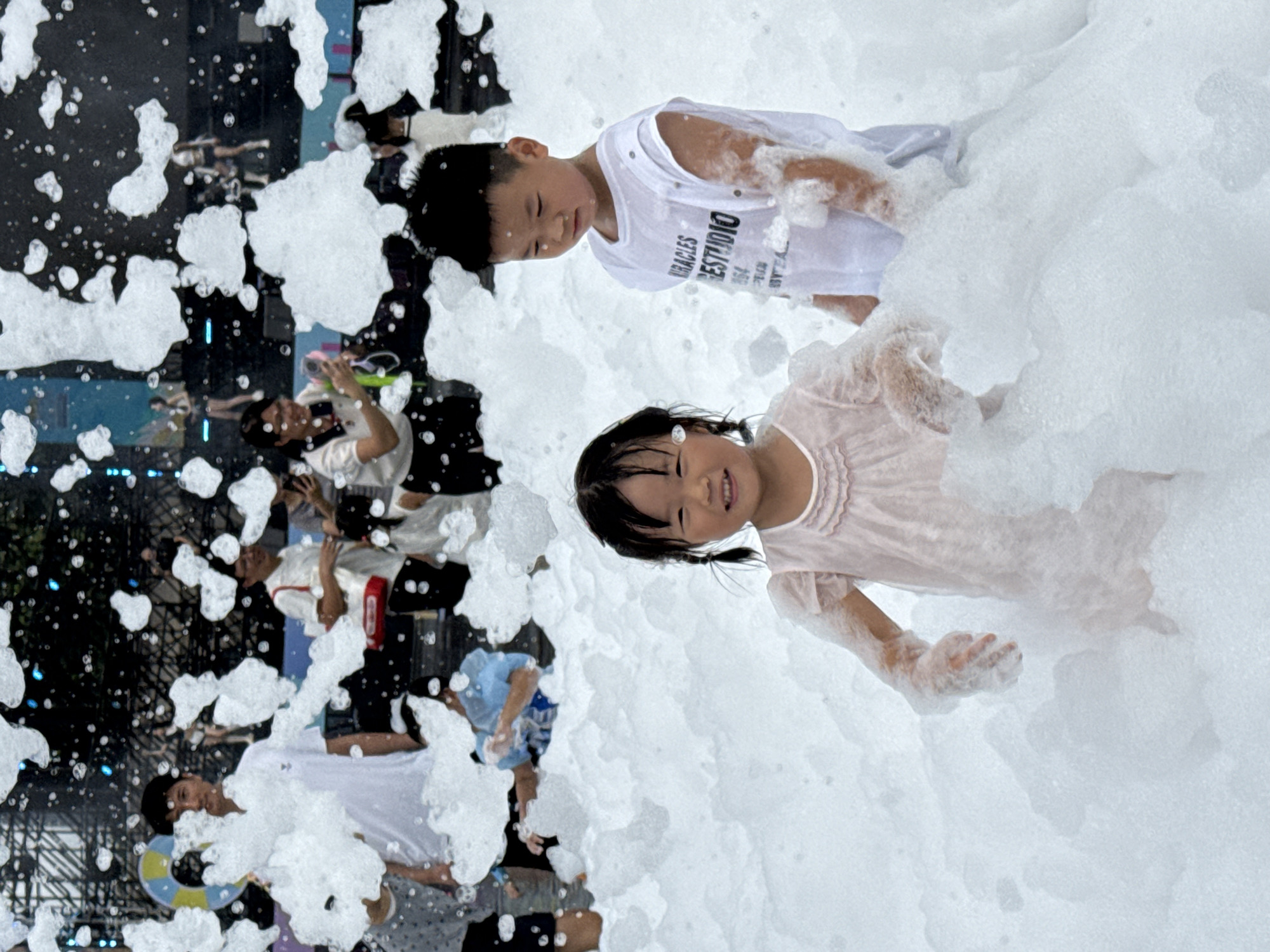 In Nanshan District, China, at an outdoor event, two young children are immersed in a large quantity of foam. The girl, on the left, is wearing a light pink dress and has pigtails. She is smiling and looking towards the camera. To her right, a boy in a white sleeveless t-shirt with text printed on it is also present, looking down. Both children's arms are covered in foam. In the background, several adults are visible, some holding objects and others looking at their phones. The environment is filled with a dense layer of white foam, obscuring the ground and some of the people. The background suggests a stage or performance area with lighting and structures.