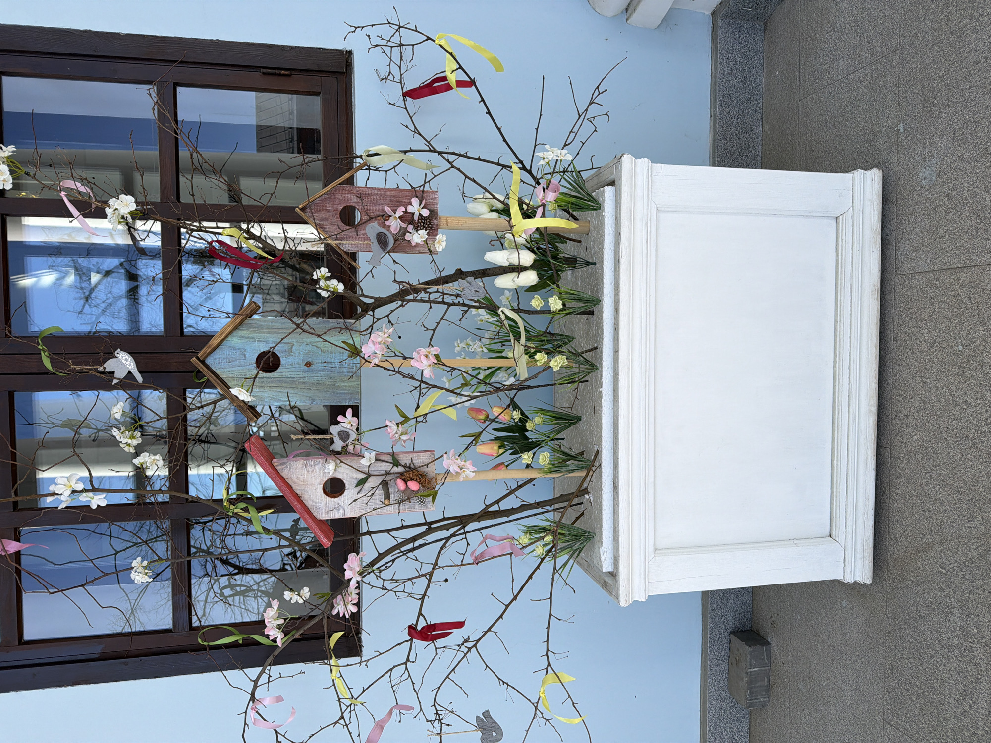 The image depicts a springtime floral arrangement in Votkinsk, Russia. The arrangement is housed in a white, rectangular planter box and consists of bare branches decorated with faux flowers, colorful ribbons, and miniature birdhouses. The birdhouses are of various designs and colors, some including decorative elements like small birds and artificial eggs. The arrangement stands outside, positioned in front of a window with a dark brown frame and pale blue walls. The reflection in the window shows bare trees and sky, suggesting early spring. The ground is paved with grey stone.