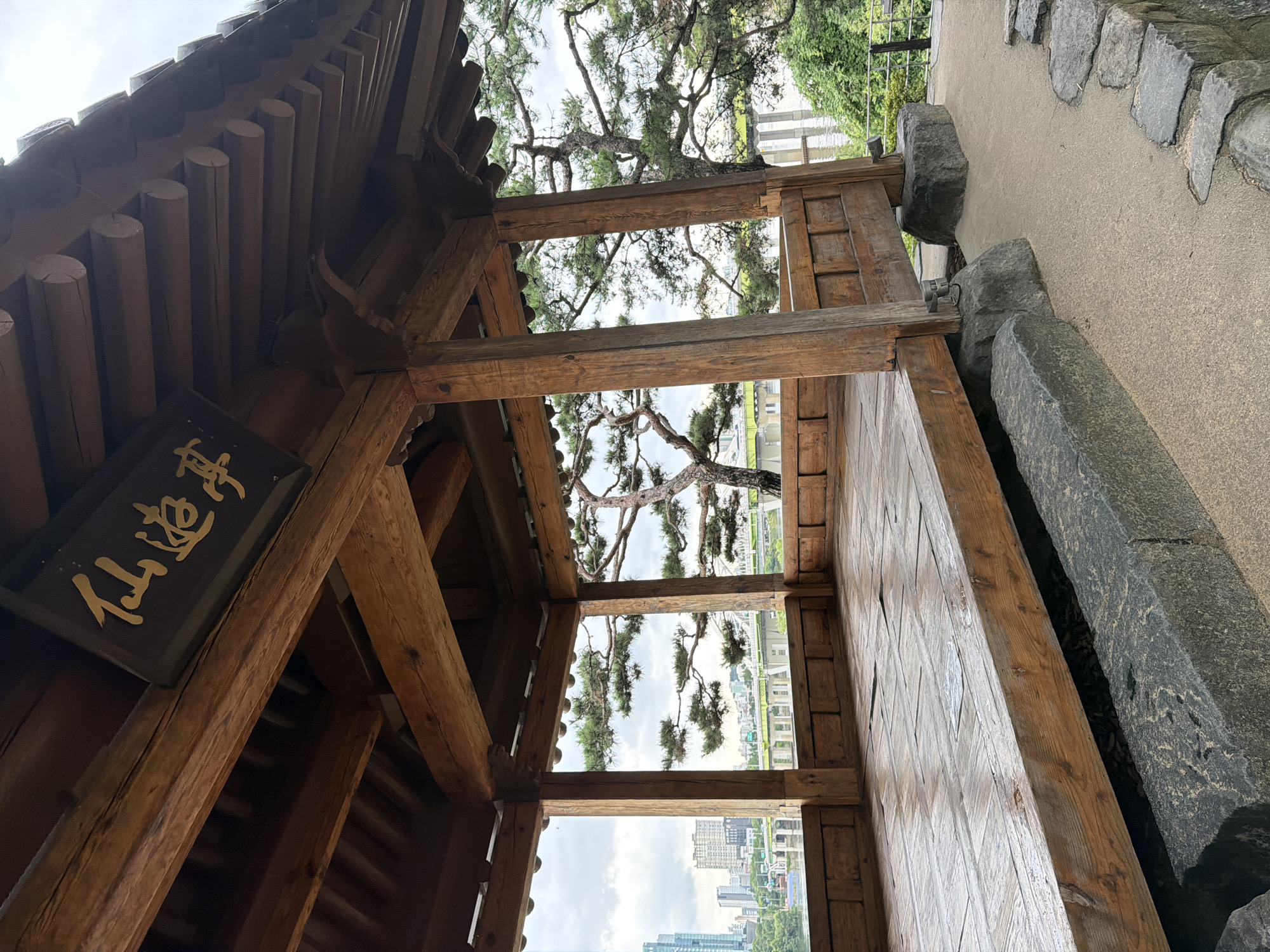 The image captures a traditional wooden pavilion with a dark wooden sign inscribed with golden Korean characters, located in Seoul, South Korea. The pavilion's wooden floor is damp, suggesting recent rain. Through the open sides of the pavilion, a cityscape is visible, featuring tall buildings and a bridge crossing a body of water. A mature pine tree with distinctive branching is also prominent in the background, adding a natural element to the urban landscape. The ground around the pavilion is covered with a light-colored sandy material and bordered by rough-hewn stones. The sky is overcast. There are no people visible in the image.