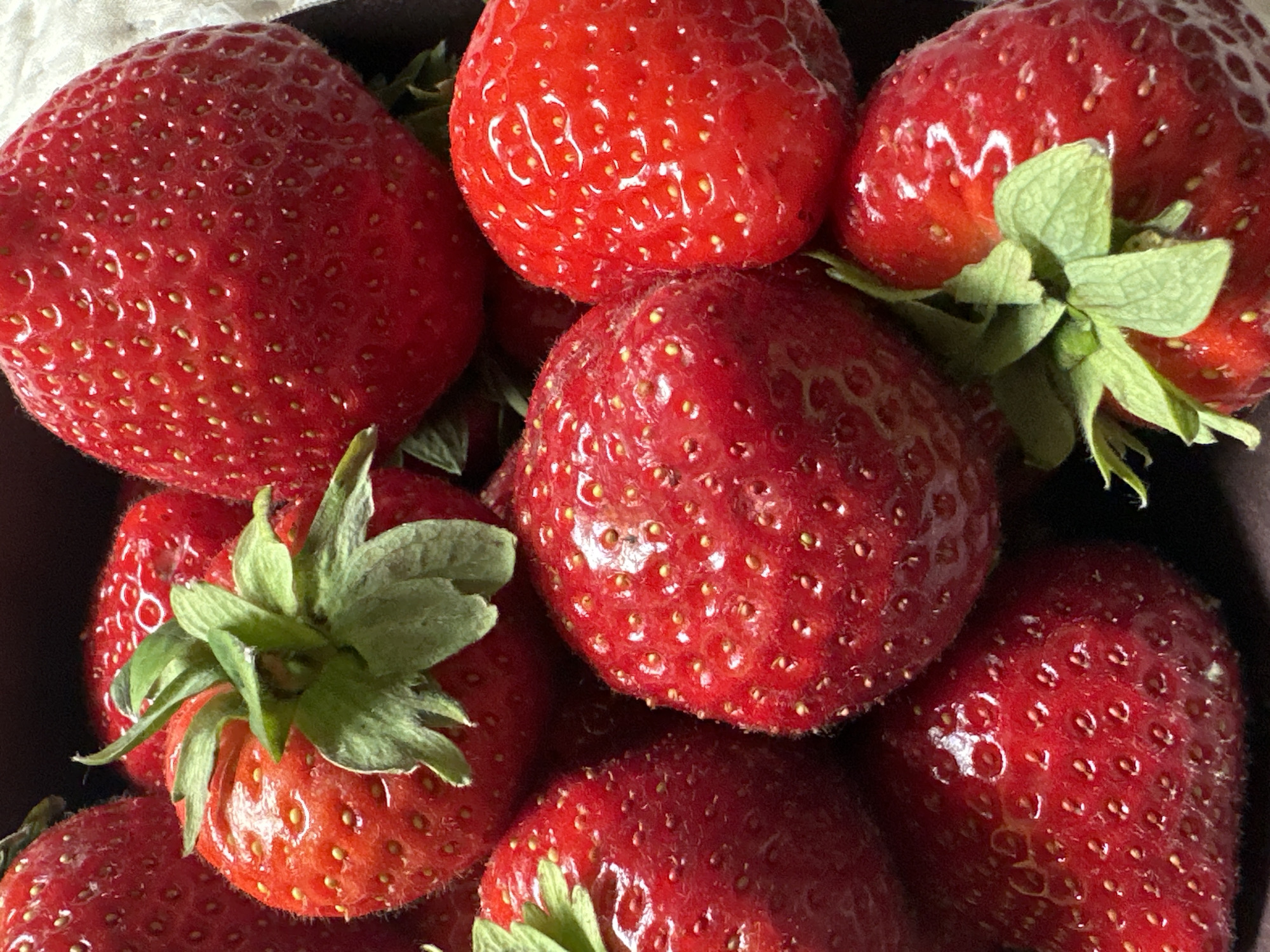 The image is a close-up, overhead shot of a collection of ripe strawberries in a dark container. The strawberries are a vibrant red, with visible seeds and green leaves at the stem. The lighting highlights the glossy texture of the fruit. There are no people or any recognizable urban or natural landmarks visible, making it impossible to determine a specific location in Tallinn, Estonia or anywhere else. The focus is solely on the strawberries.
