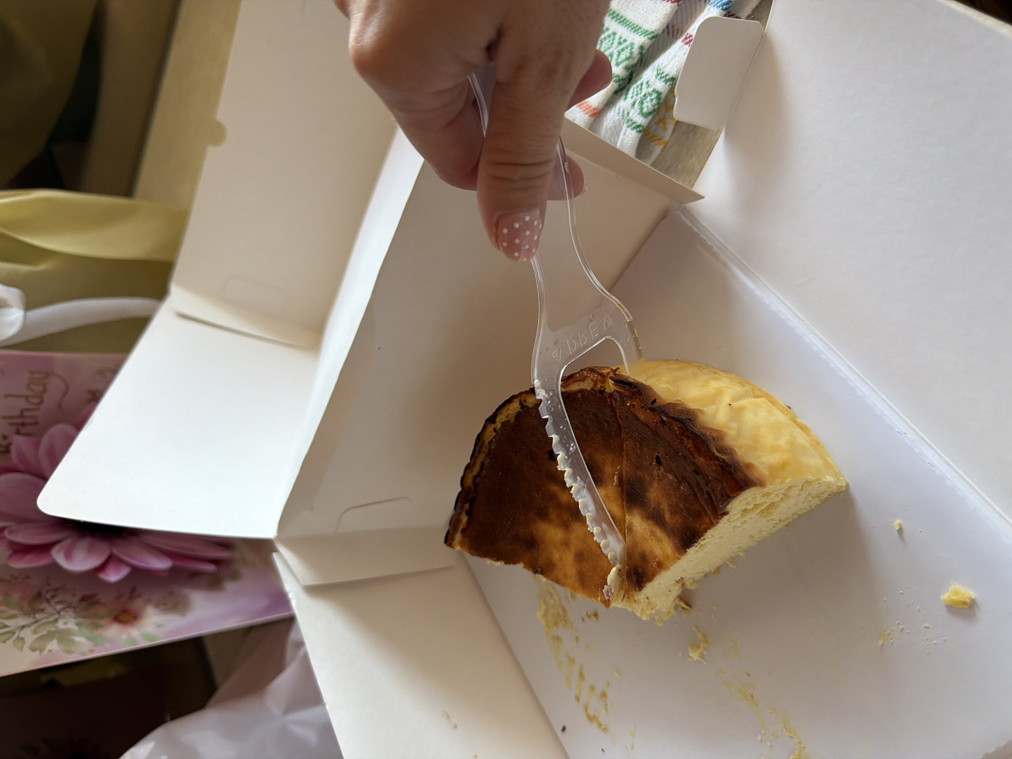 An eye-level, close-up shot captures a hand using a clear plastic cake server to cut or lift a slice of cake from a white cardboard box. The hand, belonging to an individual, features light pink fingernails decorated with white polka dots. The cake appears to be a Basque cheesecake, distinguished by its deeply caramelized, almost burnt top and a light, creamy interior, with crumbs visible on the bottom of the box.
The white cardboard cake box is partially open. To the left and below the box, a pink greeting card with floral patterns is visible, displaying the partial word "thday," suggesting a birthday celebration. A soft yellow fabric or ribbon is also present in the background on the left. On the right side, a white fabric with a green and orange patterned design can be seen. The plastic cake server has embossed text, possibly "PBE" and a recycling symbol.
The setting appears to be indoors, likely during the daytime due to bright, even lighting. The activity suggests the enjoyment of a dessert, possibly at a celebratory event. The location context for this image is Mengwi, Indonesia.