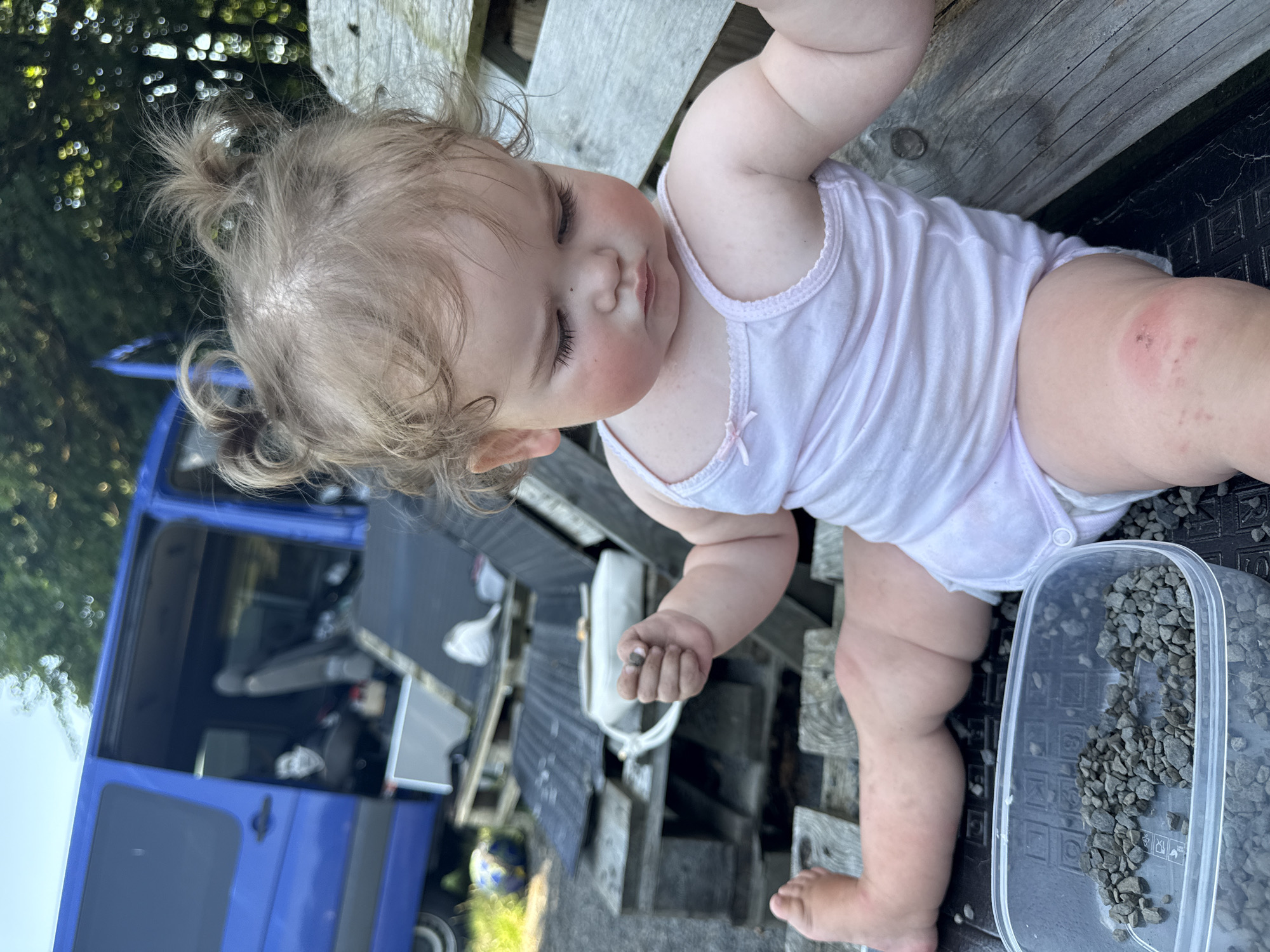 A young child is sitting on wooden pallets near a blue van, with a clear plastic container filled with small stones in front of them. The child, with light brown hair pulled into two small buns, is wearing a light pink tank top and a diaper. Their face is turned downwards, with flushed cheeks and a serious expression. One hand is raised, holding a small stone, and the other is resting on their lap. Their legs are extended, with a visible red mark on their right knee. In the background, the open door of the van reveals a dark interior, and trees can be seen beyond. The overall setting appears to be an outdoor area, possibly a yard or workshop, located in Beltershein, Germany.