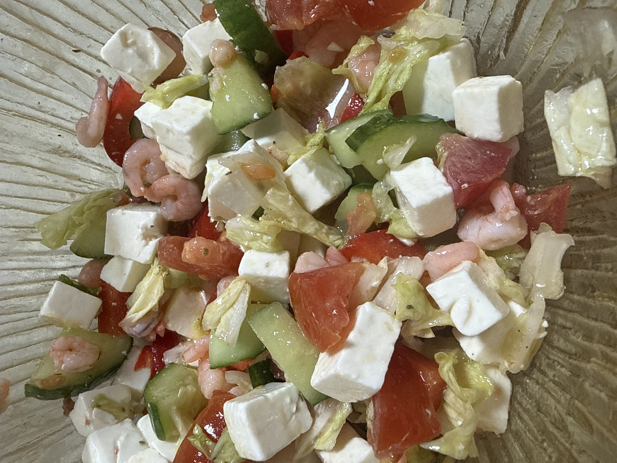 A top-down, close-up shot reveals a mixed salad in a shallow, clear glass bowl. The salad consists of diced white cheese, likely feta, mixed with chopped red tomatoes, sliced green cucumbers, small pink shrimp, and torn pieces of green lettuce. The ingredients are tossed together, appearing slightly glossy, possibly from a light dressing. The bowl's surface texture is faintly visible, with subtle ridged lines. The location is indeterminate.