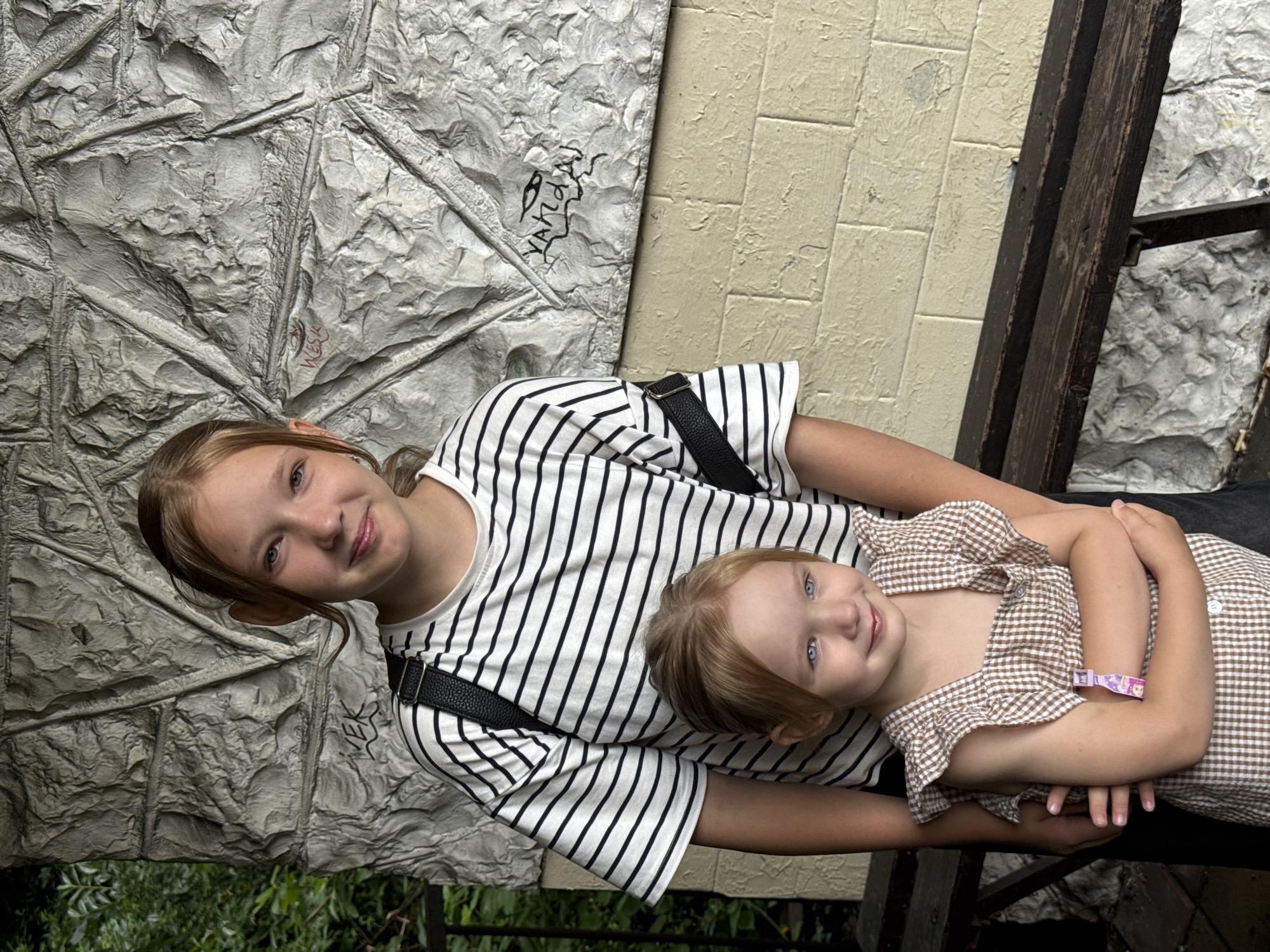 Two young females are posing for a photo outdoors, positioned in front of a textured, light-colored wall with some dark graffiti. The older girl, on the right, is wearing a black and white striped t-shirt and a black backpack strap. She has light brown hair pulled back and is smiling at the camera. The younger girl, on the left, is wearing a brown and white checkered dress with ruffled sleeves. She has light blonde hair and blue eyes and is looking slightly off-camera with a gentle smile. Both are standing close together, suggesting they are related. The background consists of the textured wall and some greenery behind a dark railing on the left side. A dark wooden bench is visible in the lower portion of the image. The overall impression is of a casual, relaxed moment captured outdoors. The context of Sochi, Russia, suggests a potentially warm climate and a public or recreational area.