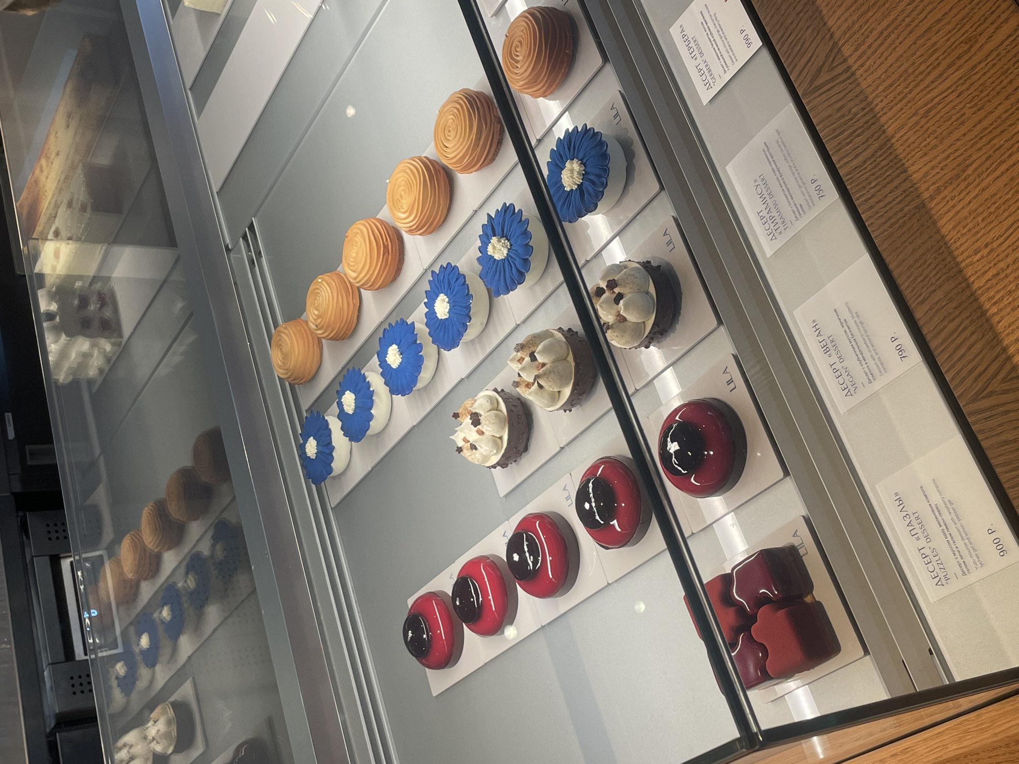 Here is a description of the image:
**Scene:** A display case in a likely upscale pastry shop, possibly in Moscow, Russia, showcasing a variety of individual desserts. The case is refrigerated, as evidenced by the slightly foggy glass.
**Subjects:** The primary subjects are the desserts themselves. From top to bottom, they appear to be:
* Round, swirled pastries (possibly meringue-based) with a golden-brown color.
* White pastries decorated with blue "flower" icing.
* Small, dark chocolate desserts topped with cream and sprinkled with toasted nuts or chocolate flakes.
* Red-glazed pastries with a dark center.
* Red-glazed desserts shaped like interlocking puzzle pieces.
**Objects:** Besides the desserts, there are:
* Small white cards placed in front of each row of desserts, likely indicating the name and price of the pastry.
* The word "LILA" is written on each of these cards.
* The prices on the cards are in Russian Rubles (P).
**Interactions/Environment:** The desserts are neatly arranged in rows within the display case. The environment appears to be clean and well-lit.