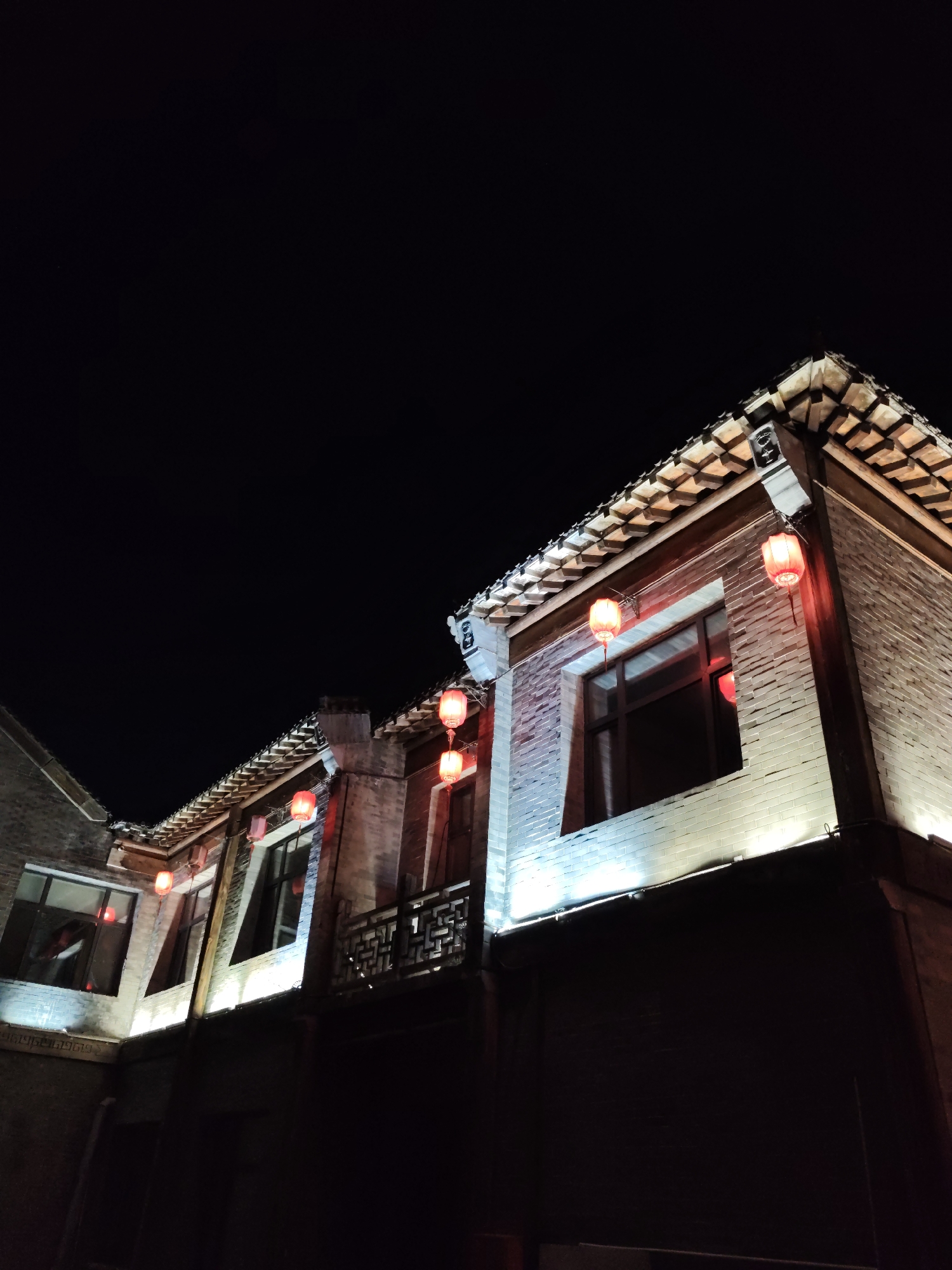 The image shows a traditional Chinese building at night, illuminated by red lanterns. The building is made of white brick and features a tiled roof. Several red lanterns hang from the eaves, casting a warm glow on the facade. Windows are visible, reflecting the light. The overall scene suggests a festive or celebratory atmosphere. The location is specified as Lin Dian Town, China. The image is a low-angle shot looking up at the building against a dark night sky.