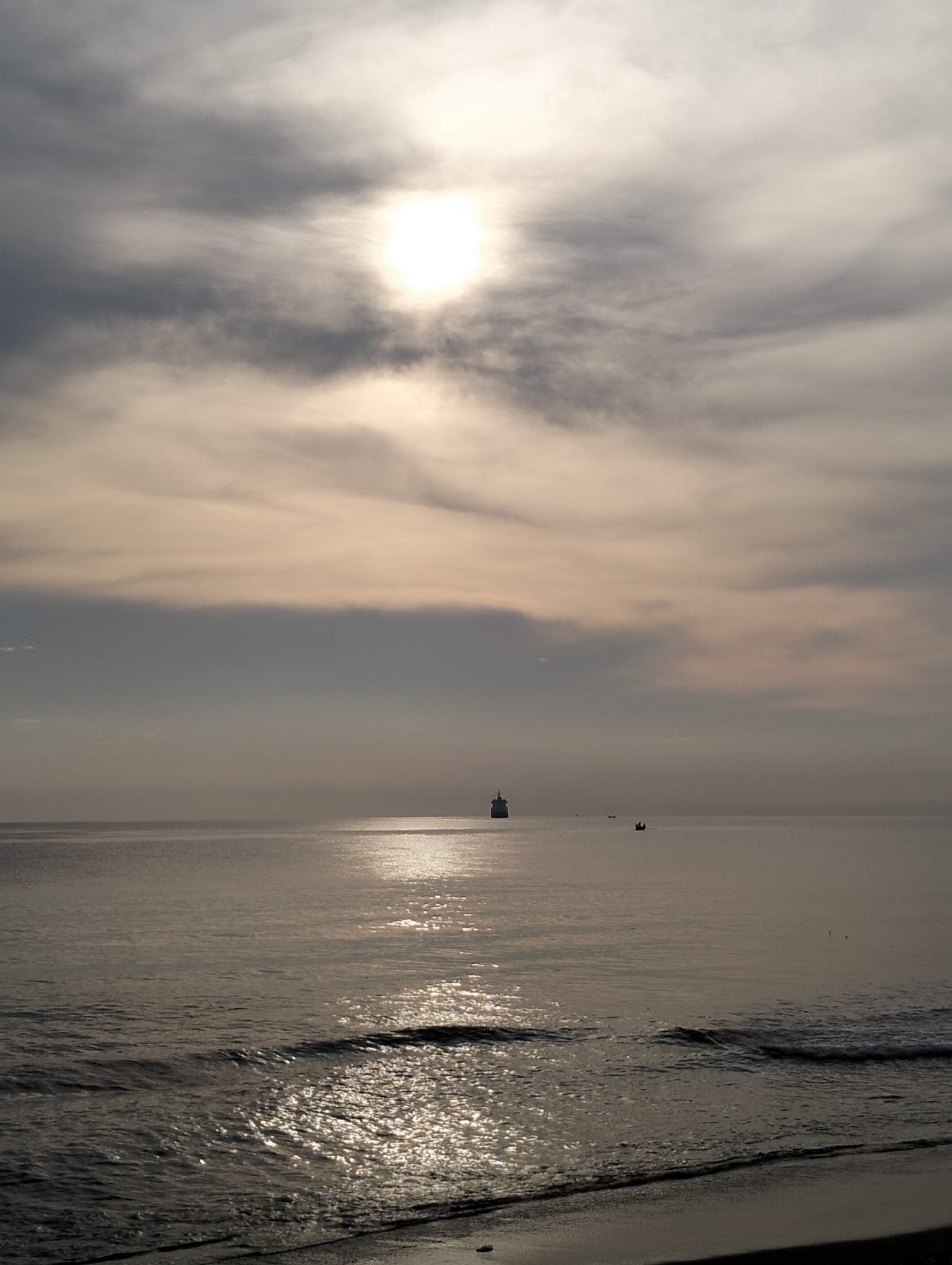 The image depicts a coastal scene likely in Lhokseumawe, Indonesia, given the prompt. The sun is high in the sky, obscured by light clouds, creating a hazy, bright light. The water is calm with ripples and reflections of the sunlight, giving it a silver sheen. There is a large ship far out at sea, along with a small boat. The foreground is a sandy beach with gentle waves breaking along the shoreline.