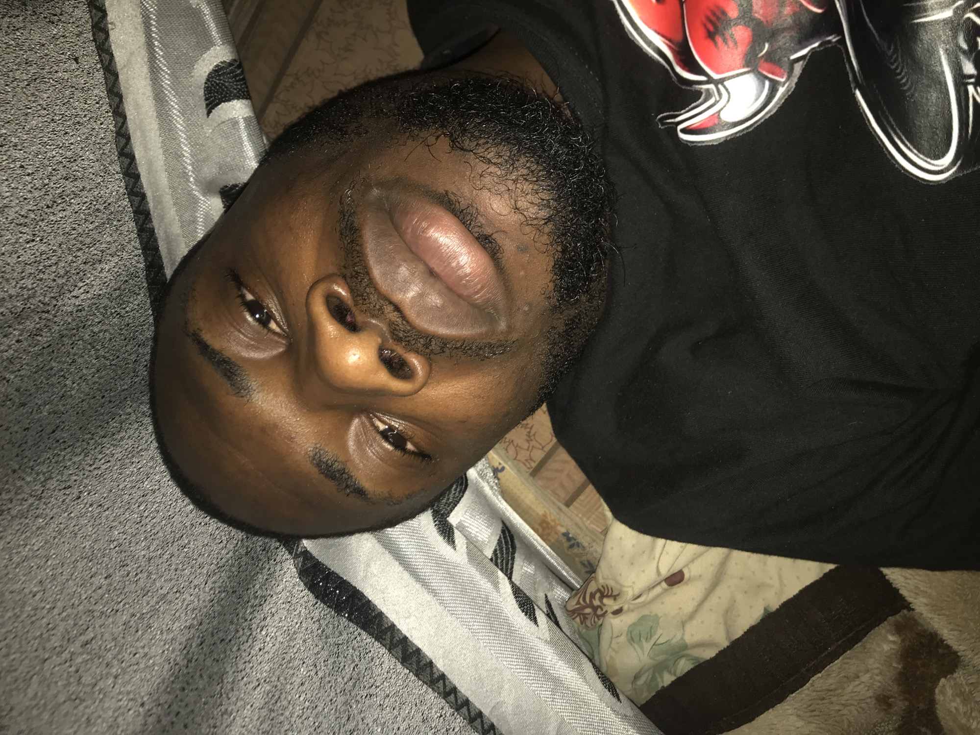 A close-up, high-angle portrait captures an adult male with dark skin, short dark hair, and a full, short beard, lying in an indoor setting. He is looking directly into the camera, with slightly bloodshot eyes and full lips. The subject is wearing a black t-shirt, partially revealing a red and white graphic design on the right side of his chest, resembling a horned, cartoon-like figure. The background consists of various textiles, suggesting a bed or couch. To the upper left, a textured grey fabric is visible, transitioning into a lighter striped fabric with dark geometric patterns. Below and to the right of the subject, a light-colored sheet or pillowcase with green and brown floral patterns is present. A beige, fuzzy blanket is also visible in the lower left portion of the frame. The lighting is soft and even. The scene takes place in an unspecified indoor domestic environment within the city of Unknown, Unknown.