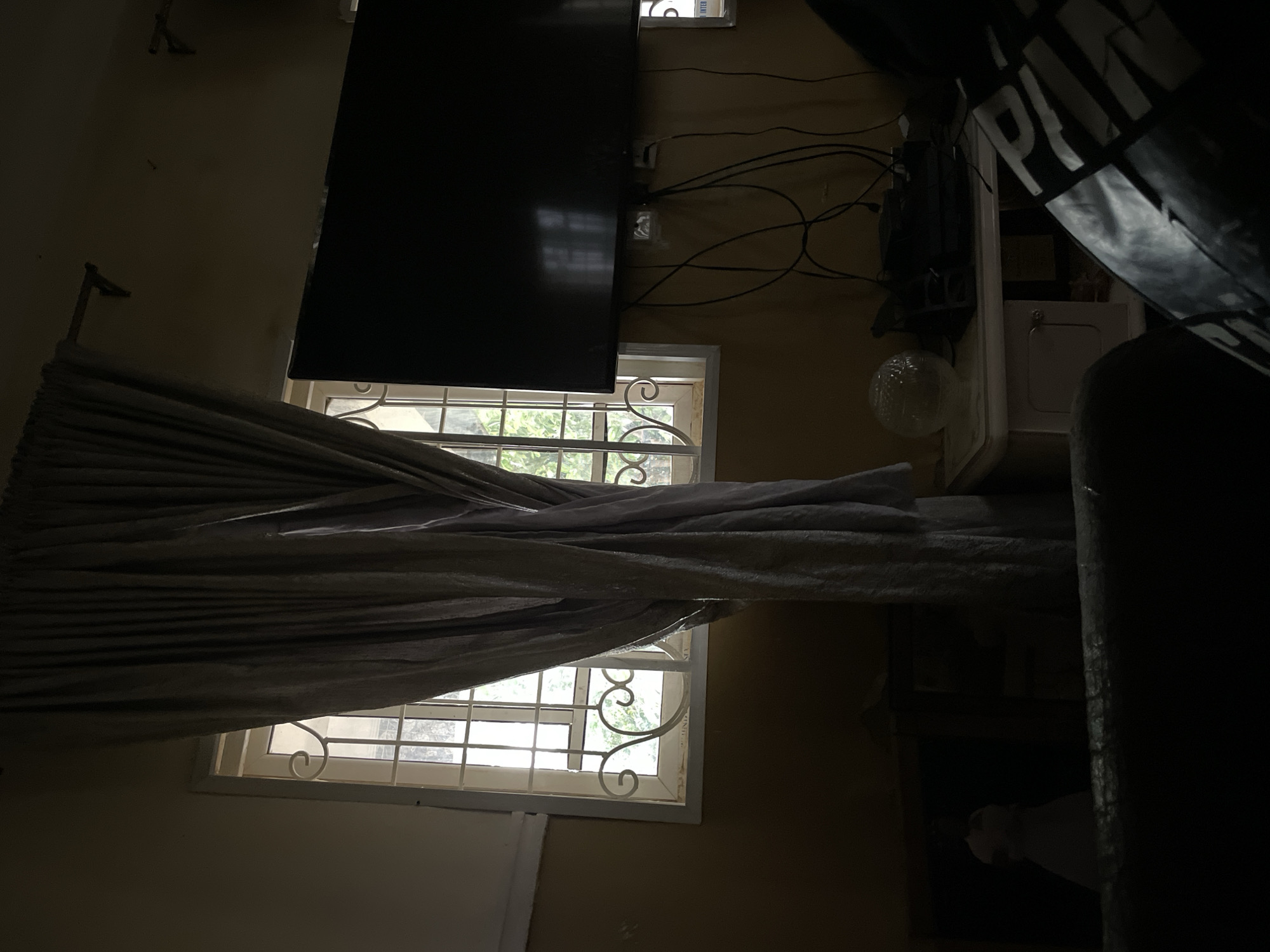 The interior scene appears to be a darkened room in Rafin Bauna, Nigeria. A television is mounted on the wall above a window with decorative bars, partially obscured by a curtain. Wires from the television are visible. The room is dimly lit, making it difficult to discern further details about the furnishings or inhabitants. A partial view of what could be a piece of furniture is seen on the right edge.