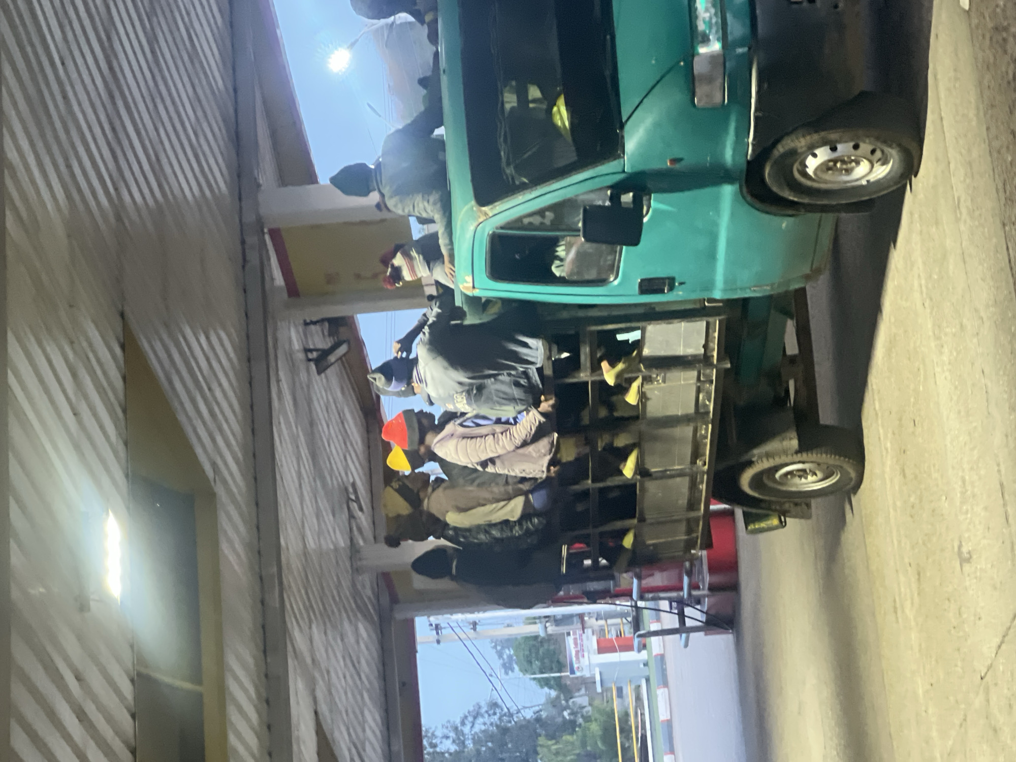 The image depicts a crowded green truck, likely a light-duty or medium-duty model, transporting multiple people. The truck, with an open cargo bed, has numerous individuals, mostly men, riding in the back. Some are standing, others appear to be seated or bracing themselves within the cargo area. One person is also visible near the passenger side of the truck's cabin.
The scene appears to be set on a road or an open area. To the right of the truck, a building with a distinct corrugated metal wall is visible, reflecting a strong, directional light. In the distance, on the left side of the image, trees and other structures can be seen under a clear sky, suggesting an outdoor, possibly semi-urban or rural environment in Gura Top, Nigeria.
The activity taking place is the transportation of people, likely workers, in the back of the truck. This is a common sight in many regions.
Notable details include the time of day, which appears to be either early morning or late afternoon/dusk. A bright light source, possibly the sun or a powerful artificial light, is prominently visible in the upper-left corner of the image (when oriented correctly), casting strong shadows on the corrugated wall and illuminating parts of the scene. The sky in the background has the soft, diffused light characteristic of dawn or twilight. The individuals are dressed in casual attire, with some wearing hats or caps. Within the truck's cargo bed, some yellow or brightly colored items, possibly safety gear or personal belongings, are visible among the passengers. No discernible text is visible on any signs or vehicles in the image.