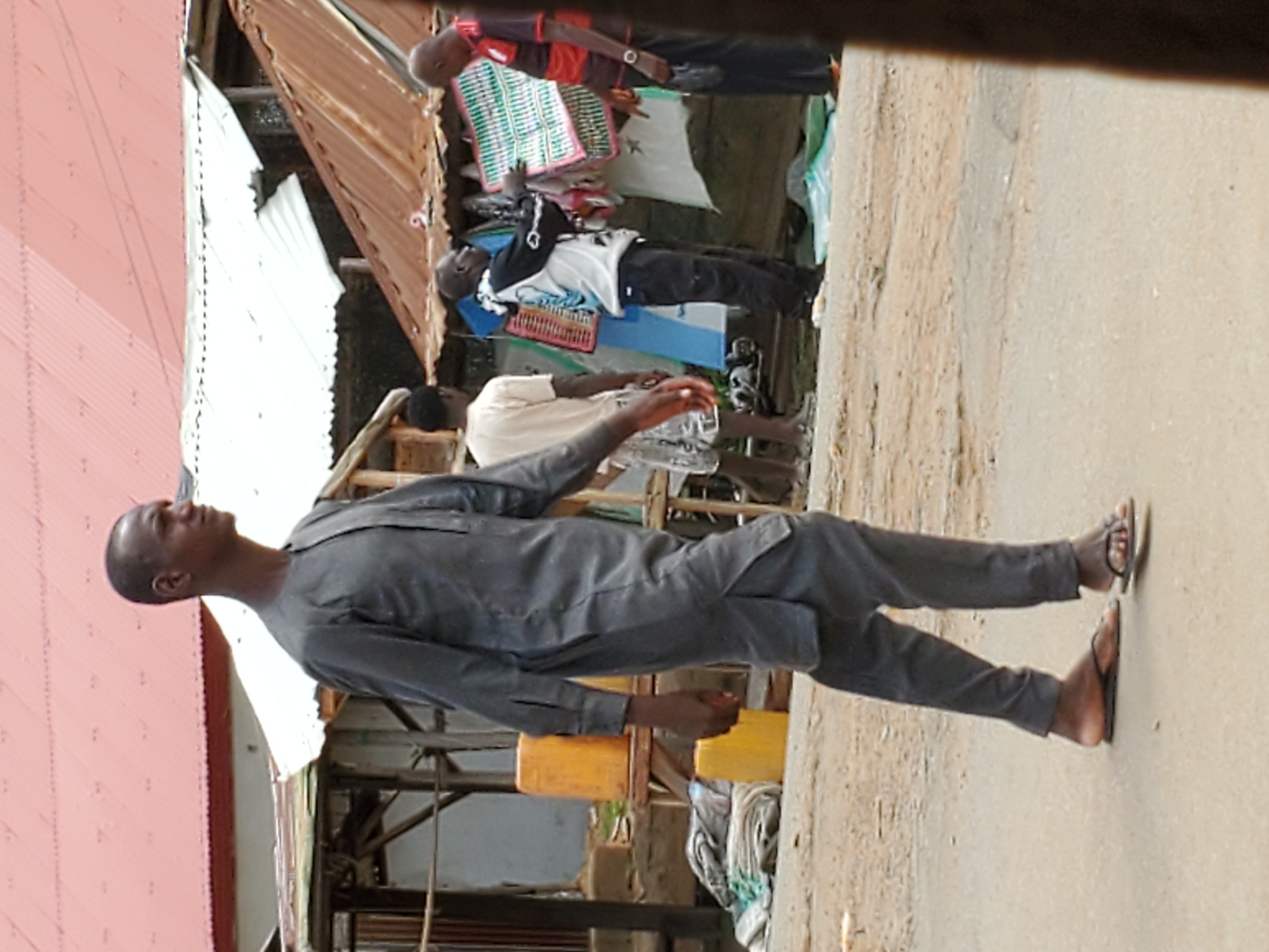 The image depicts a man in a gray tunic and pants walking on a paved surface in Mararaba, Nigeria. He is wearing sandals. Several other people are visible in the background standing near a makeshift storefront with metal roofing and various goods displayed. A yellow plastic container is visible near the man. The environment appears to be a street scene with buildings on one side.