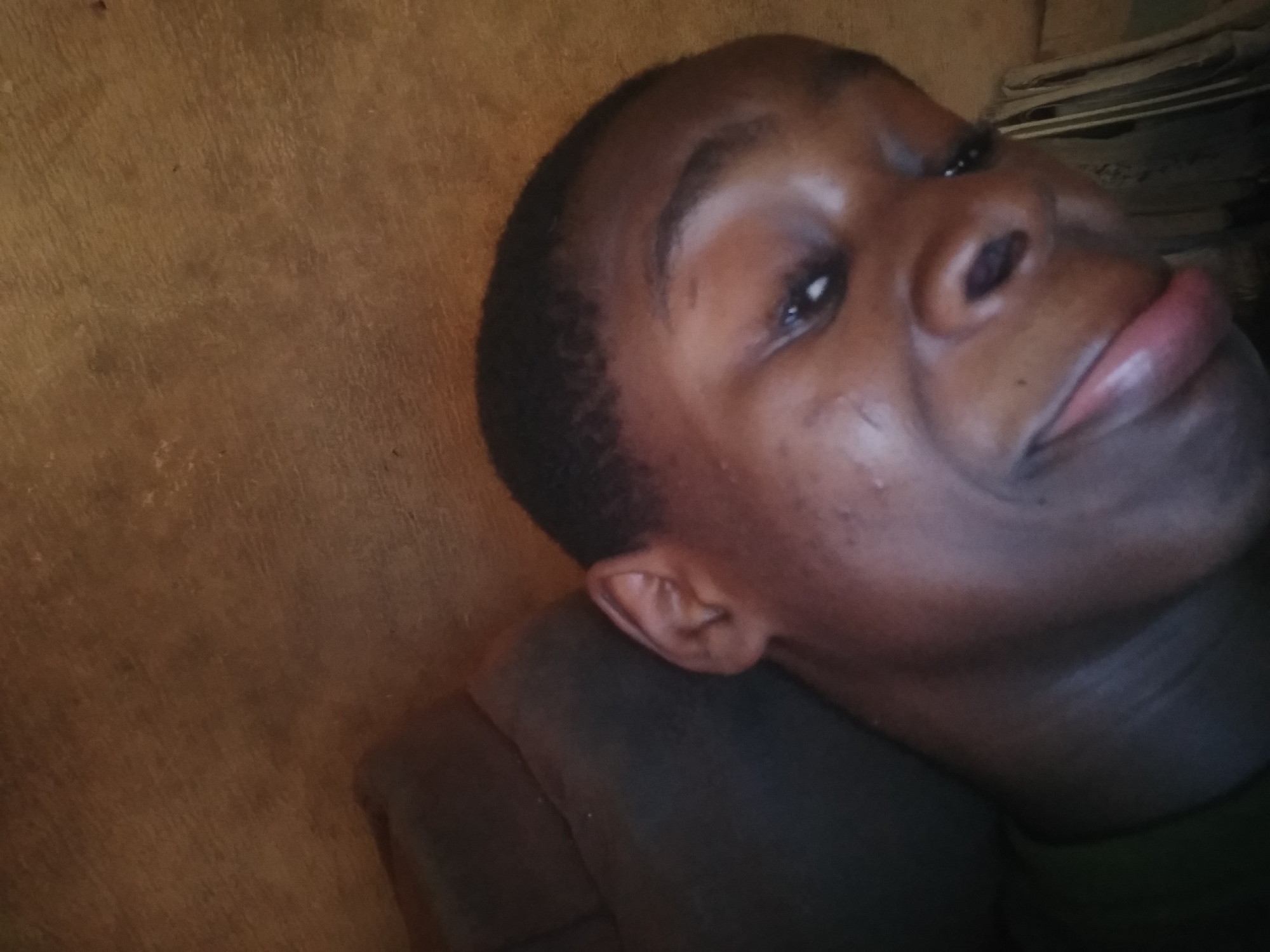 A close-up, angled portrait of a young, dark-skinned person with short, dark hair. The person is resting on a dark, soft surface, possibly a pillow or cushion. The background includes a textured brown wall on the left and a blurry stack of items on the right. The location, Shangisha, Nigeria, is not discernible from the visual information in the image.