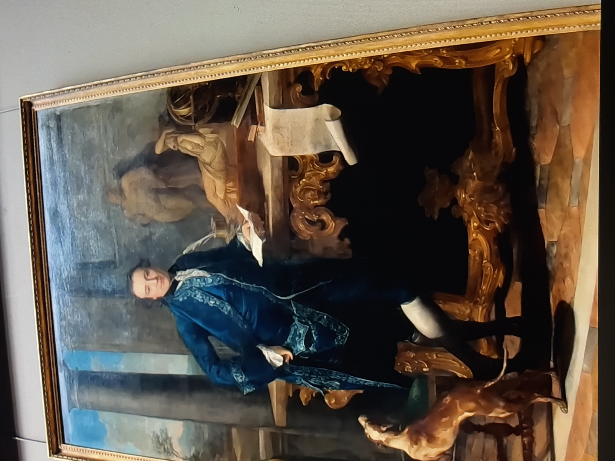 A full-length oil painting depicts a man in a blue velvet suit with embroidered trim standing next to a wooden desk. He wears white stockings and black shoes, and his hand rests on a book. The desk is ornately carved and features a globe, a scroll of paper, and a plaster statue. Behind the man, two large columns frame a statue of a reclining figure. In the foreground, a dog stands on its hind legs, looking up at the man. The painting is framed in gold and hangs on a wall. The location is Unknown, Unknown.