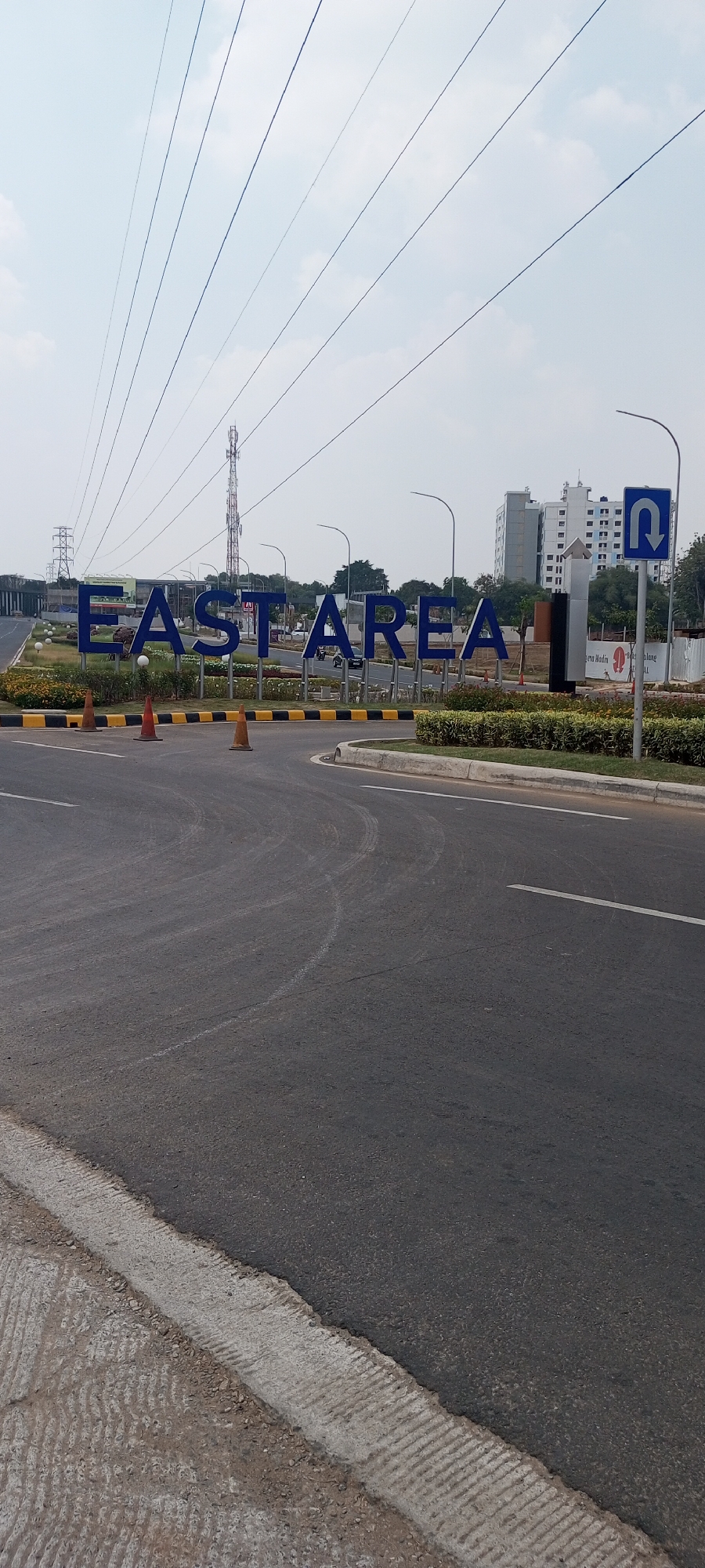 The image captures the entrance to an area with large, blue, three-dimensional letters spelling "EAST AREA". These letters are situated behind a road and a landscaped area with green bushes and white decorative spheres. Orange traffic cones are placed on the road in front of the sign. In the background, there is a cellular tower, a tall white building, and other structures under construction. A blue street sign with a white U-turn arrow is visible on the right side of the image. The sky is overcast. The scene appears to be an outdoor environment, likely a road leading into a commercial or residential development. The specific location is identified as Rawa Banteng, Indonesia.